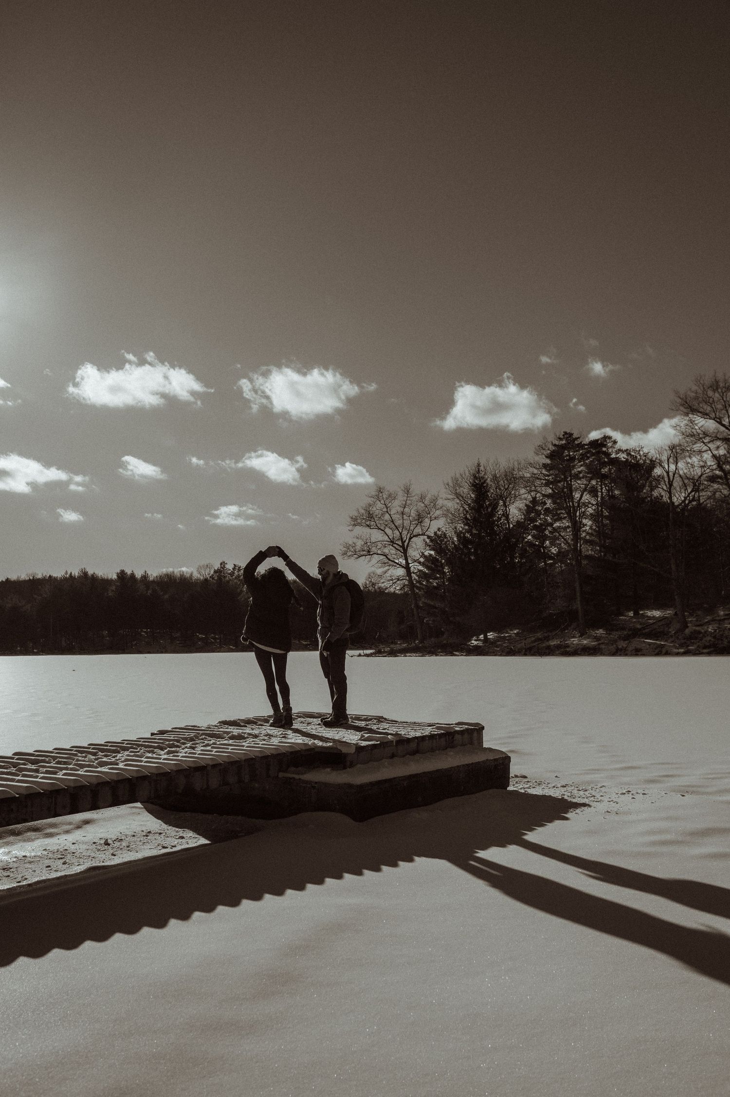 Two silhouettes stand on a dock overlooking a frozen lake in black and white.