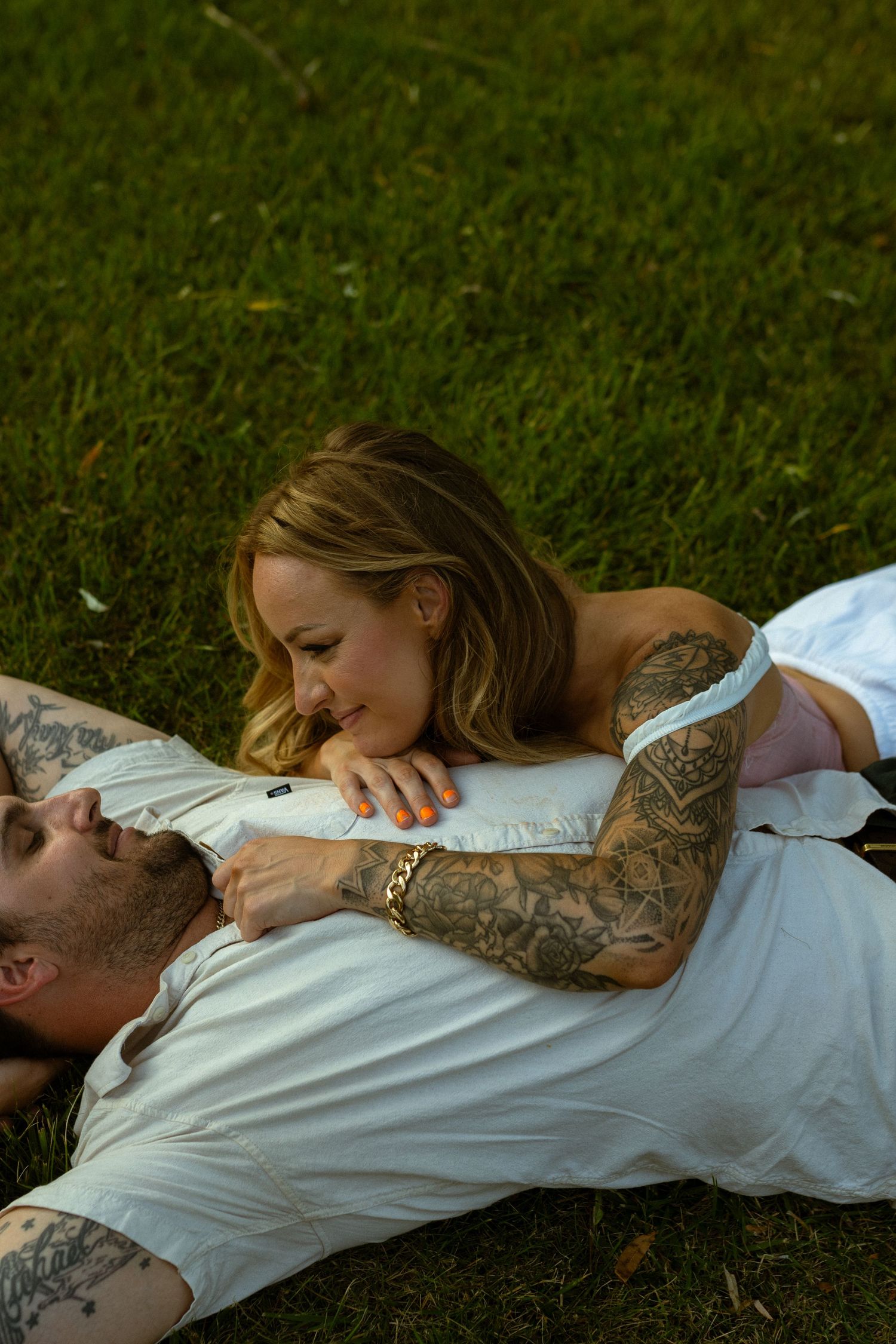 A couple lying on grass sharing tender moments with visible arm tattoos.