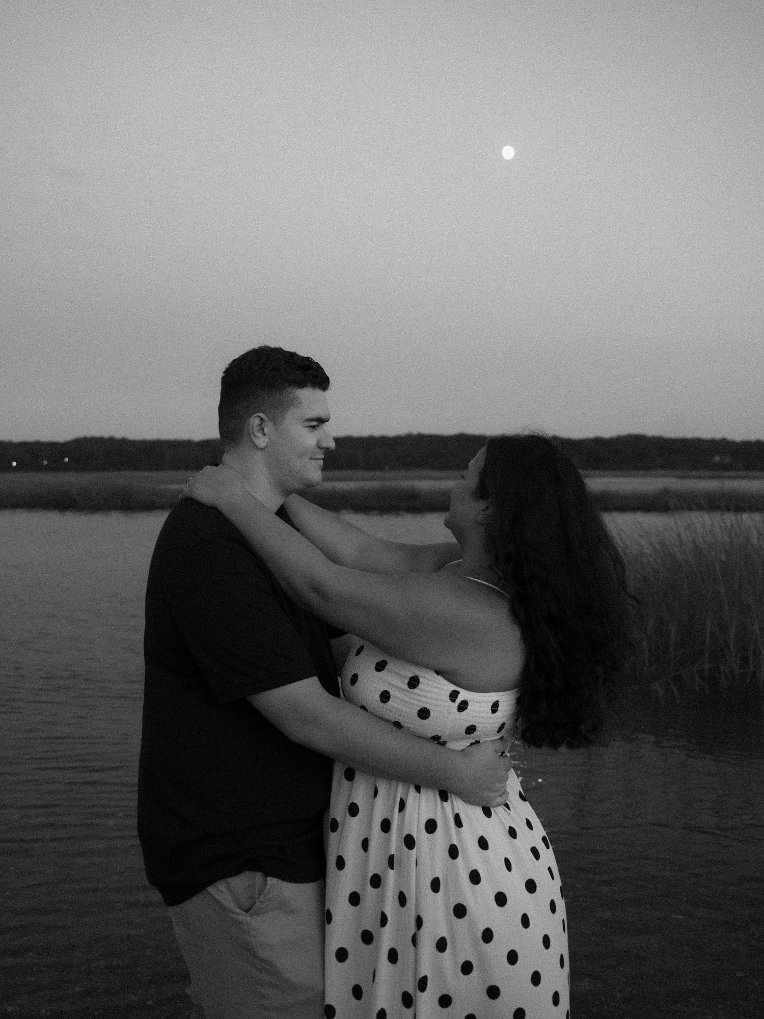 Black and white photo of a couple embracing romantically by a lake at dusk with a full moon overhead.