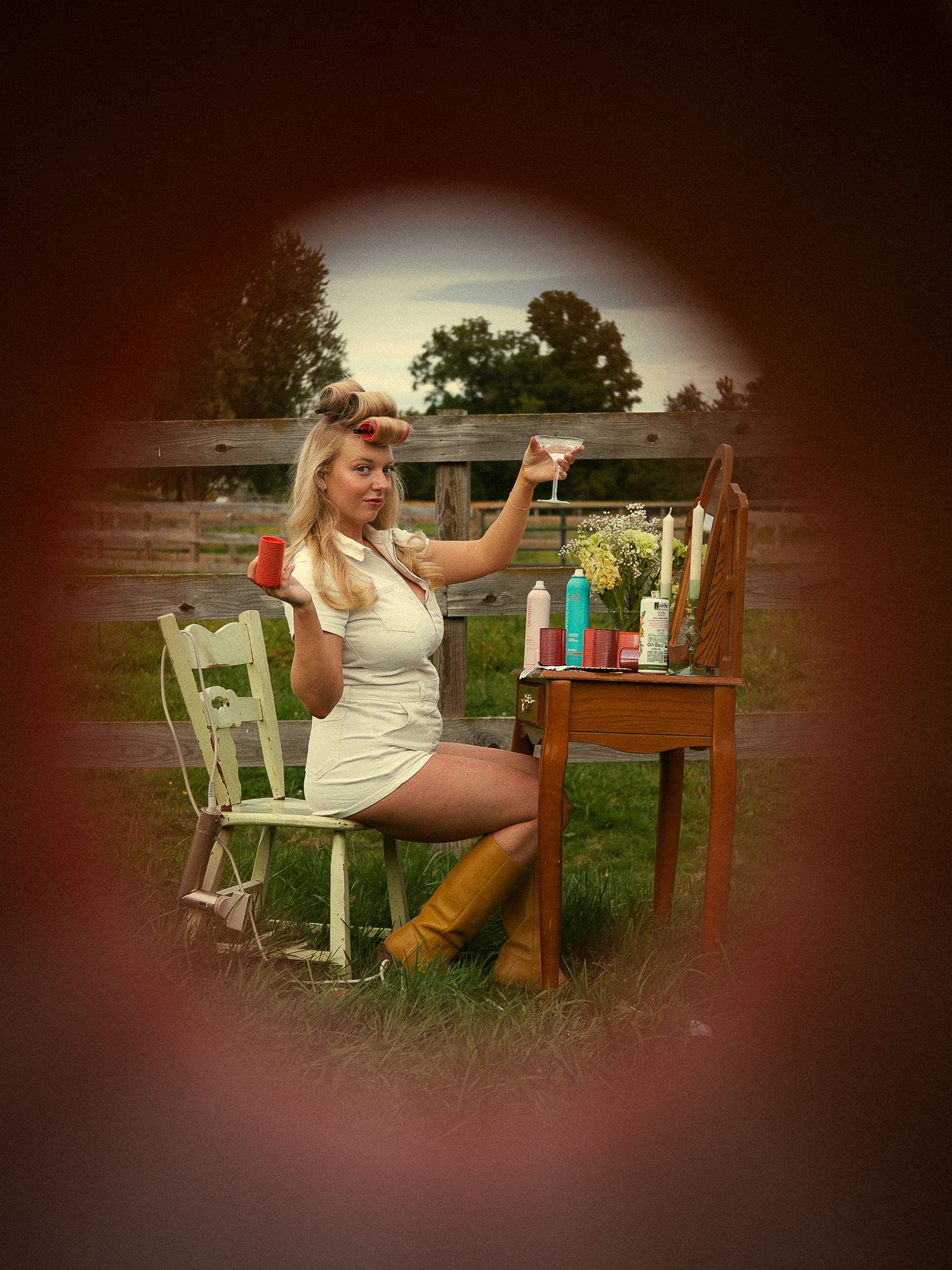 Vintage pin-up style blonde woman in white dress and yellow boots raising a glass outdoors near a rustic fence.