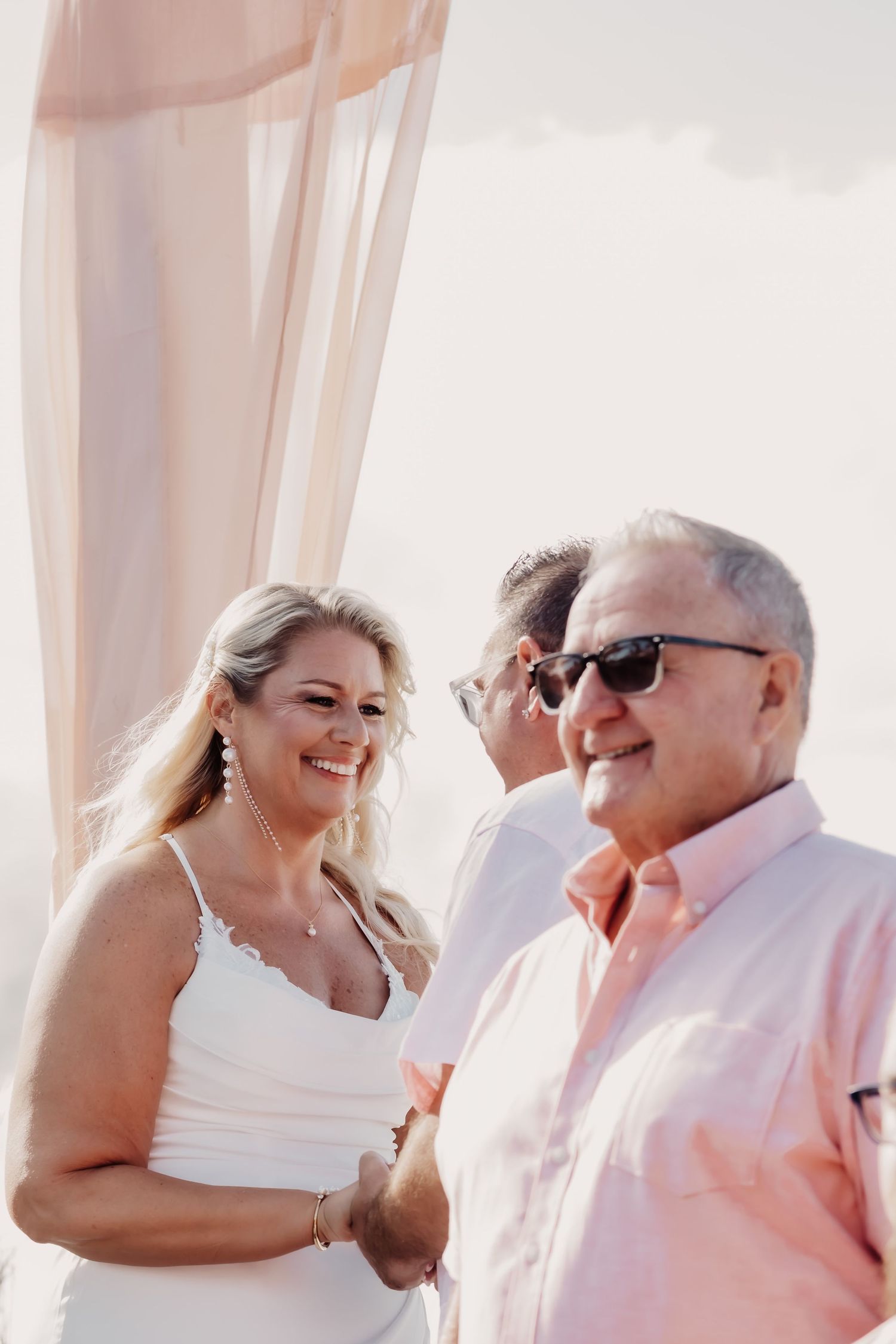 Candid wedding moments captured on a beach with light pink curtains flowing in the background.