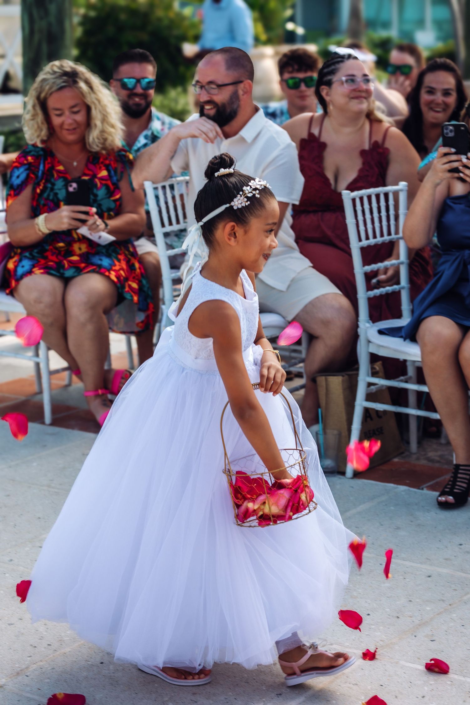 Flower girl in white tulle dress spreading rose petals during wedding ceremony.