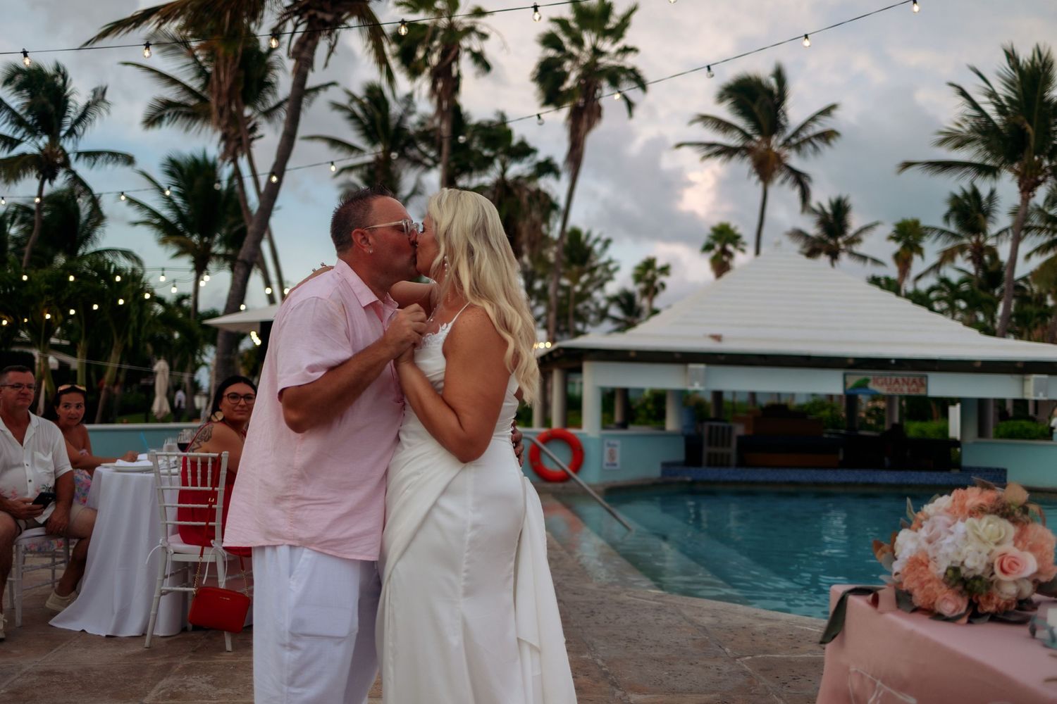 Romantic poolside wedding reception at a tropical resort with palm trees and string lights at sunset.