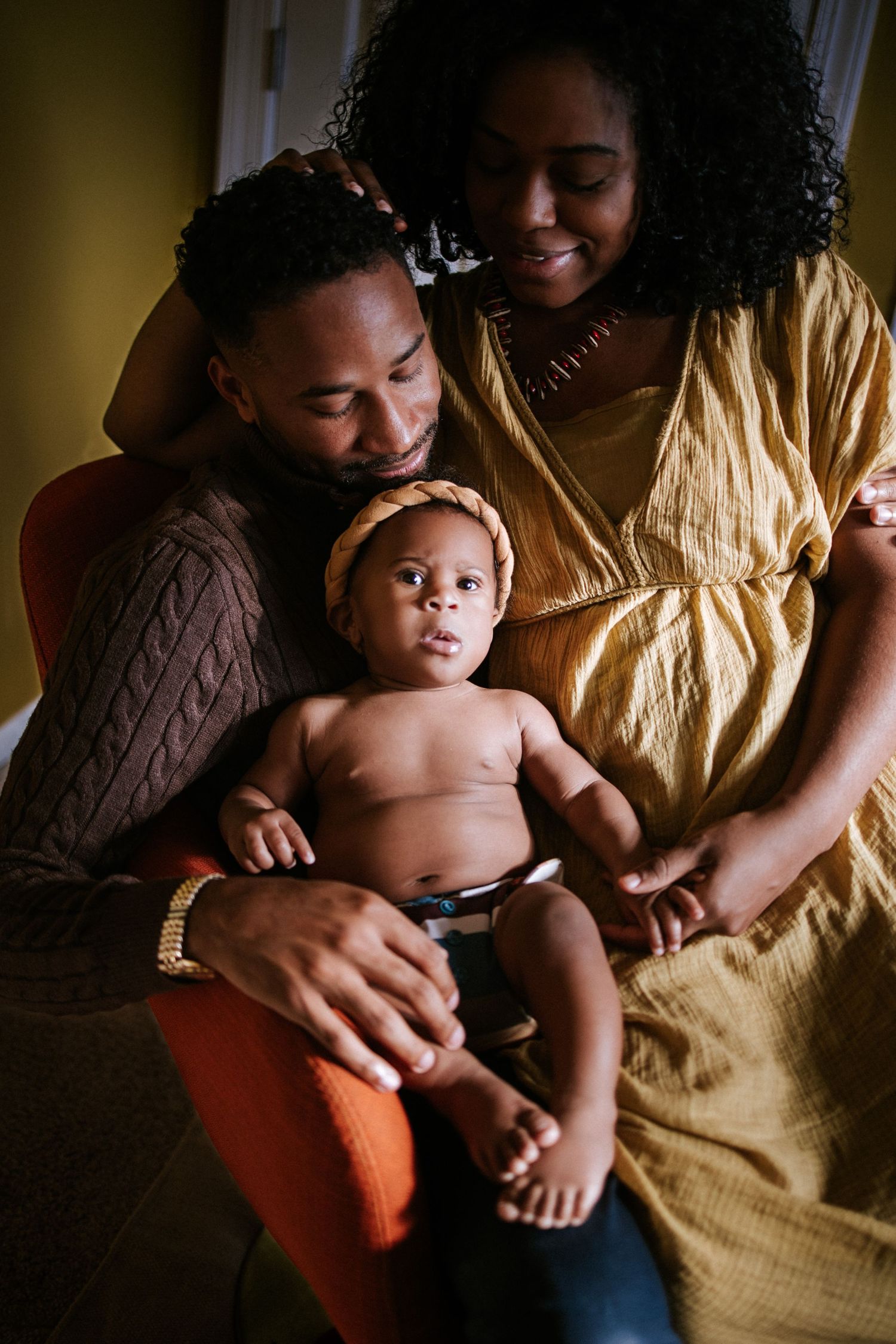 Loving family moment with baby wearing white lace against yellow backdrop.