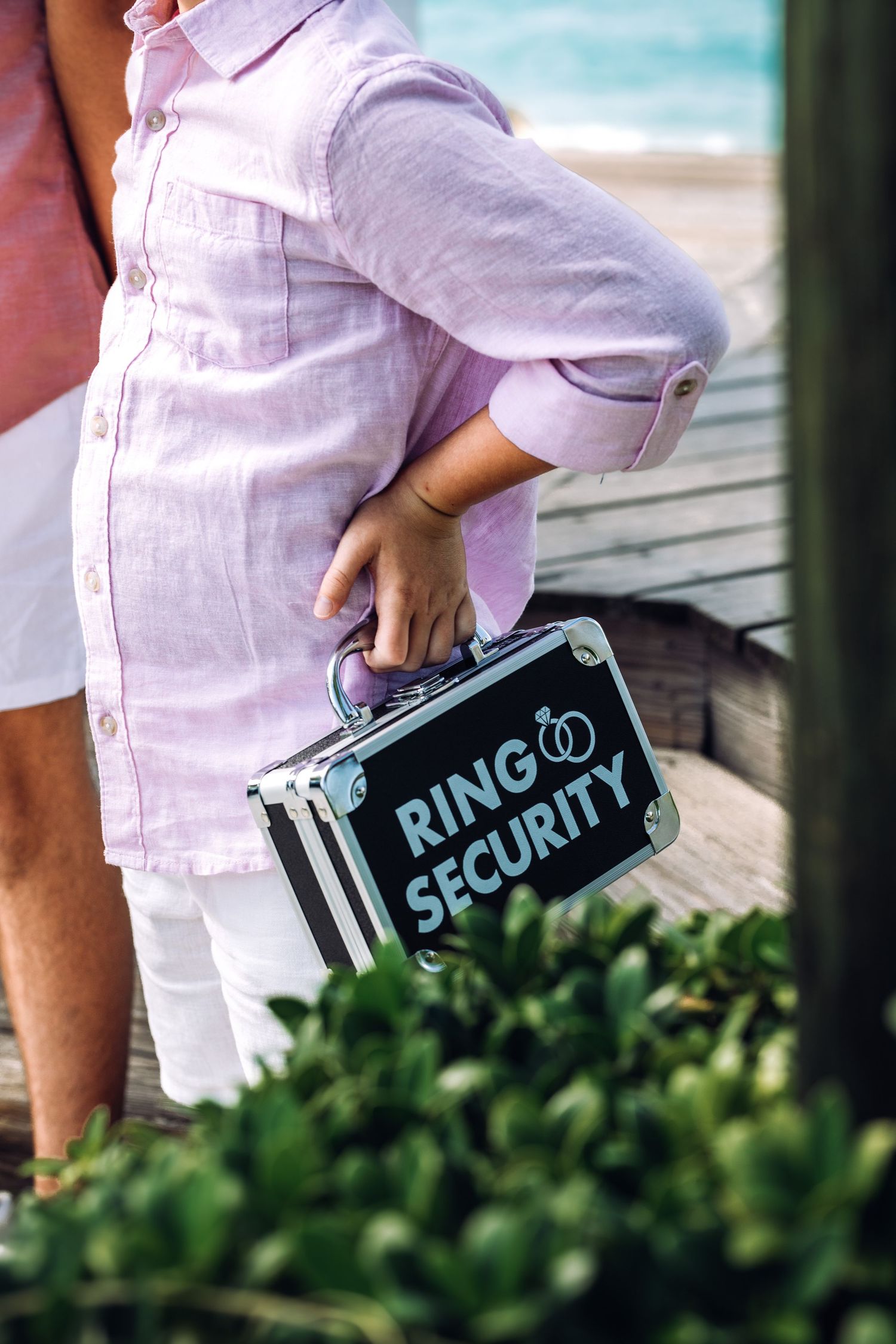 A small black sign reading 'Ring Security' is held against a garden backdrop for a beach side destination wedding.