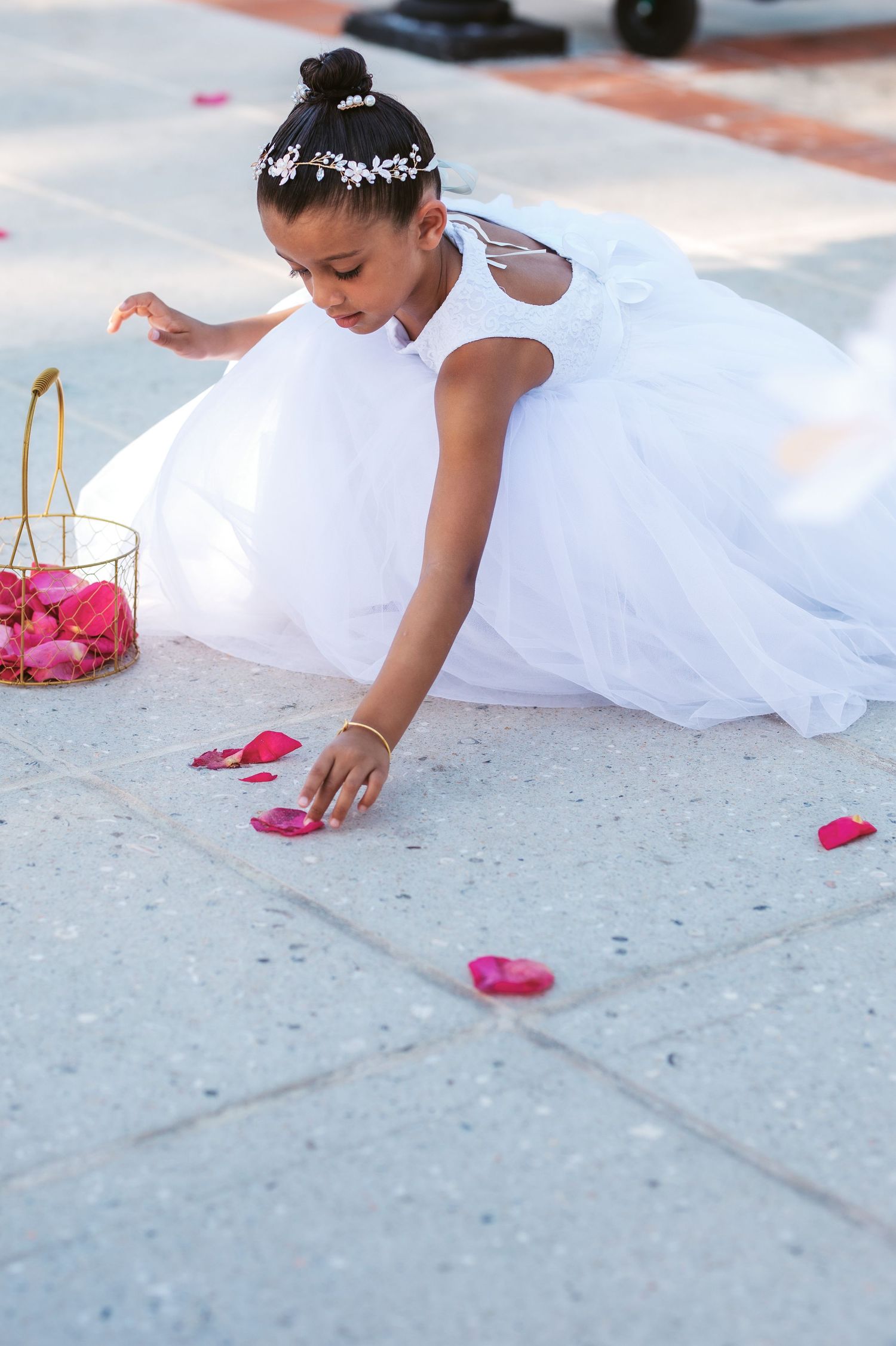 A flower girl in a white dress scatters pink rose petals on a concrete pathway during a wedding ceremony.