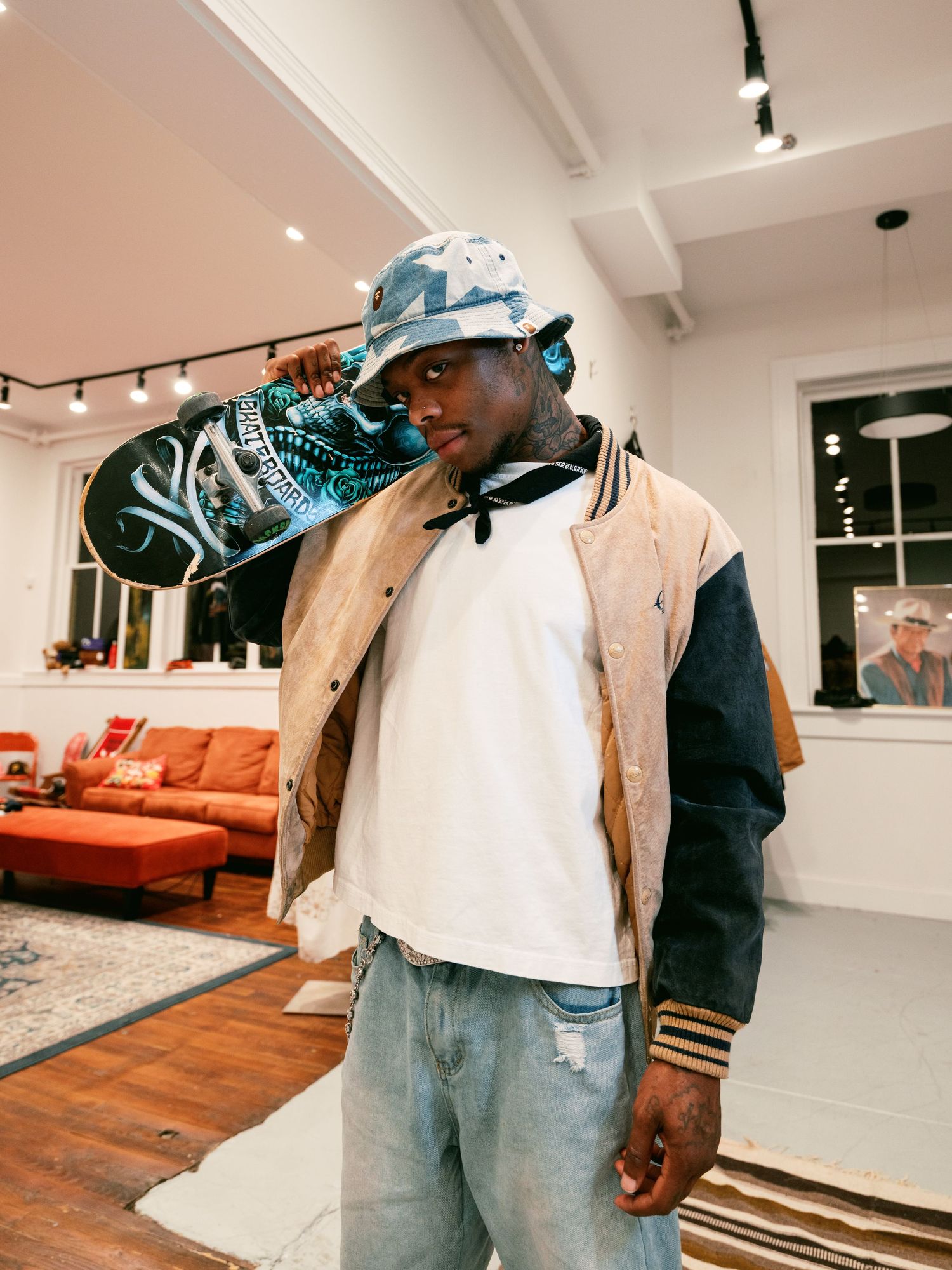 Young man in bucket hat and varsity jacket holds skateboard on shoulder indoors.