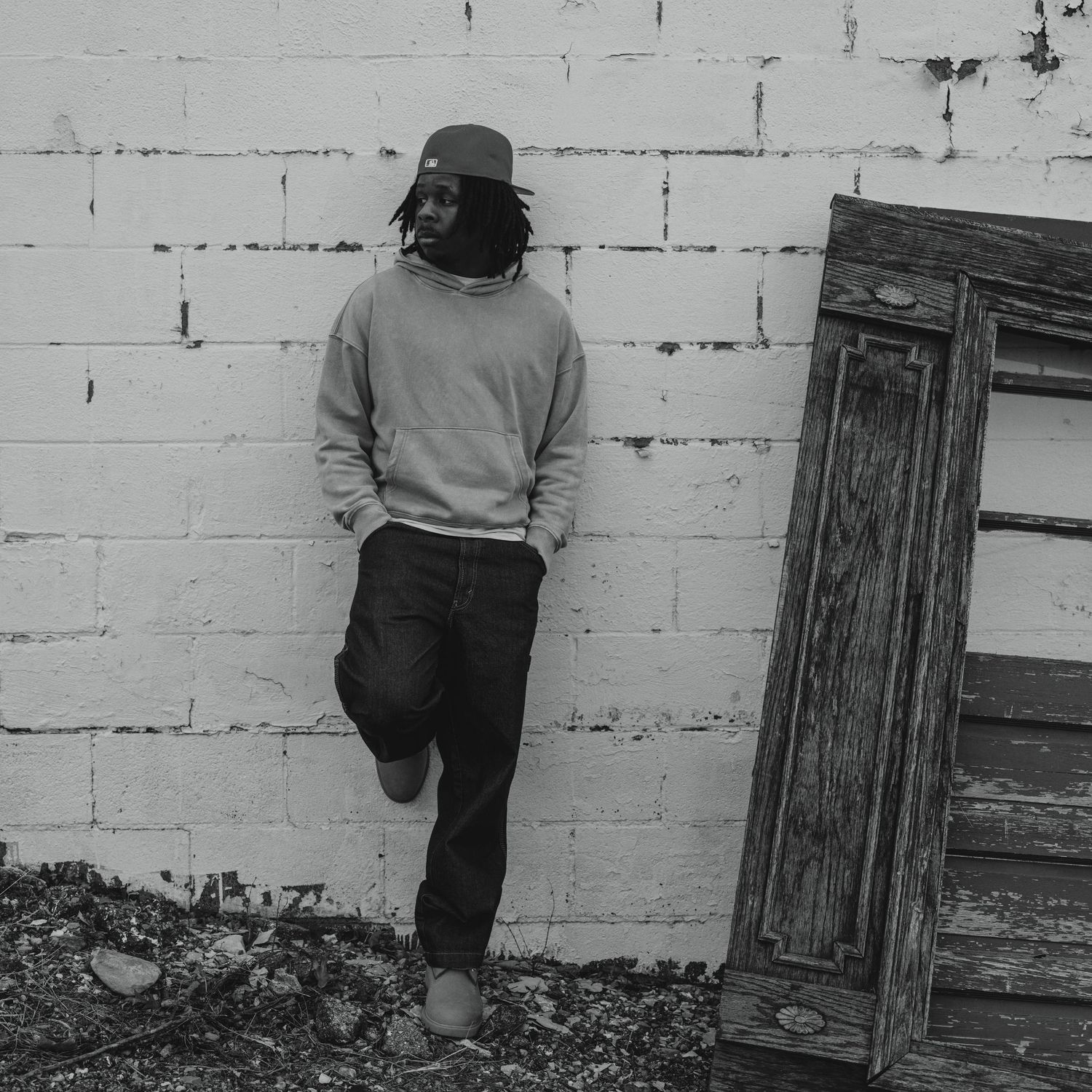 A young man in a gray sweatshirt and black pants leans against a white brick wall in a black and white photo.