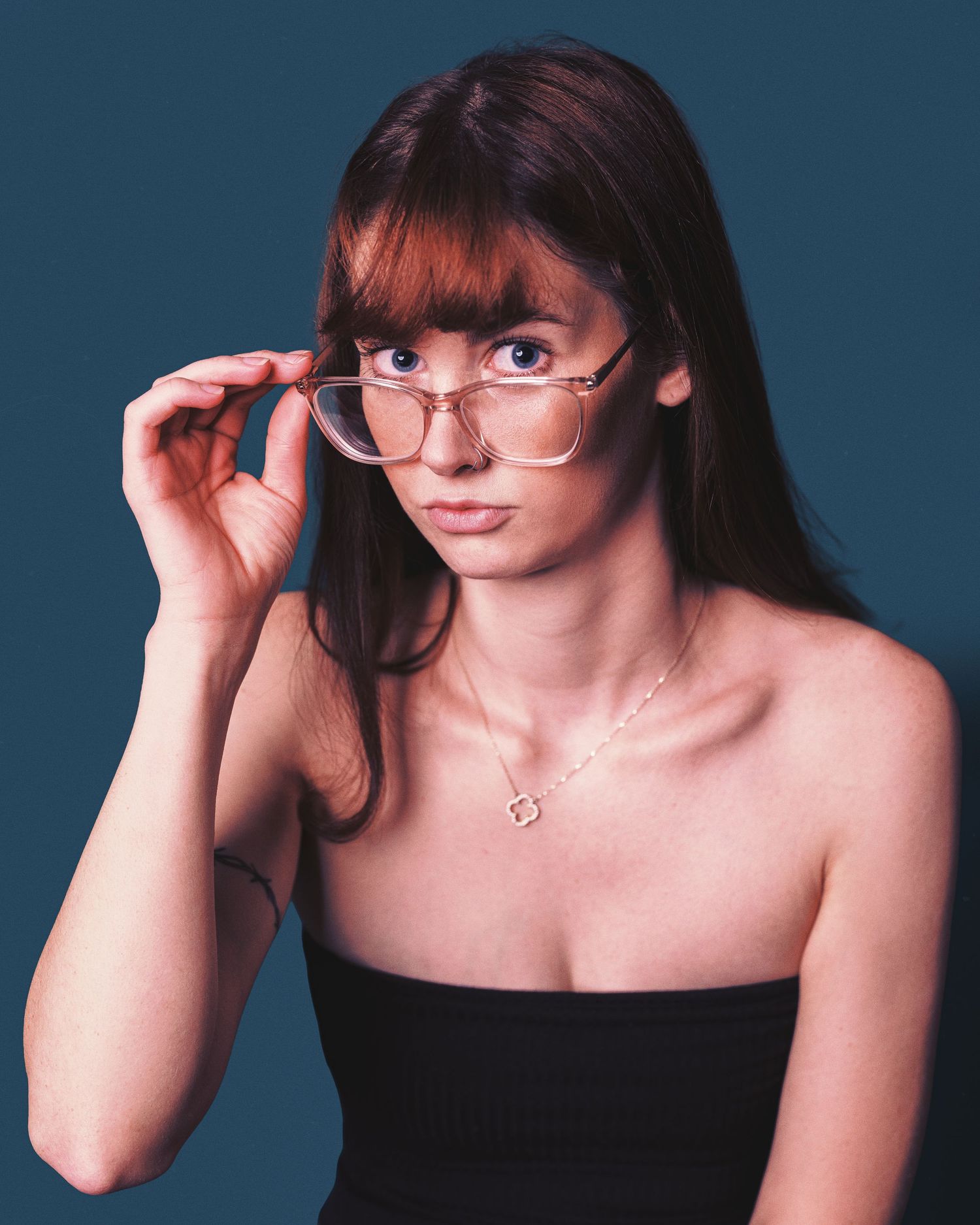 A portrait of someone wearing a black strapless top and wire-rimmed glasses against a dark blue background.