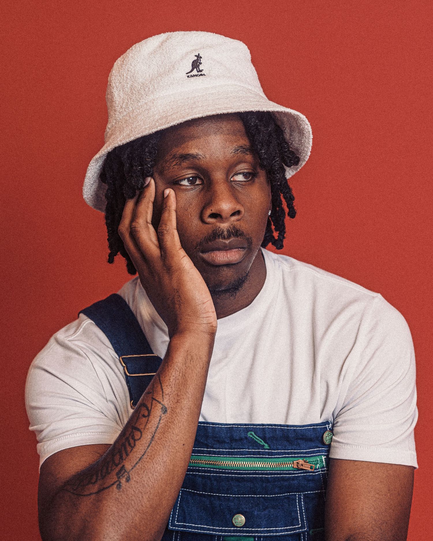 Studio portraits against red backdrop featuring someone in white t-shirt, overalls and bucket hat.