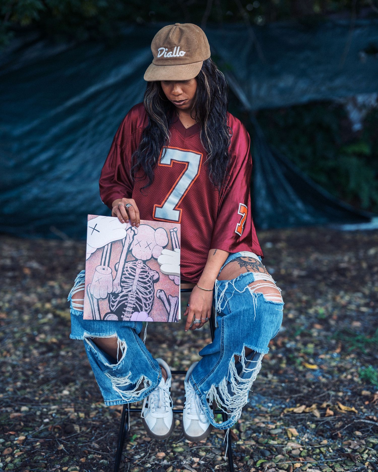 Woman in red jersey #7, ripped jeans, and tan cap holds artwork while sitting outdoors near a blue tarp.