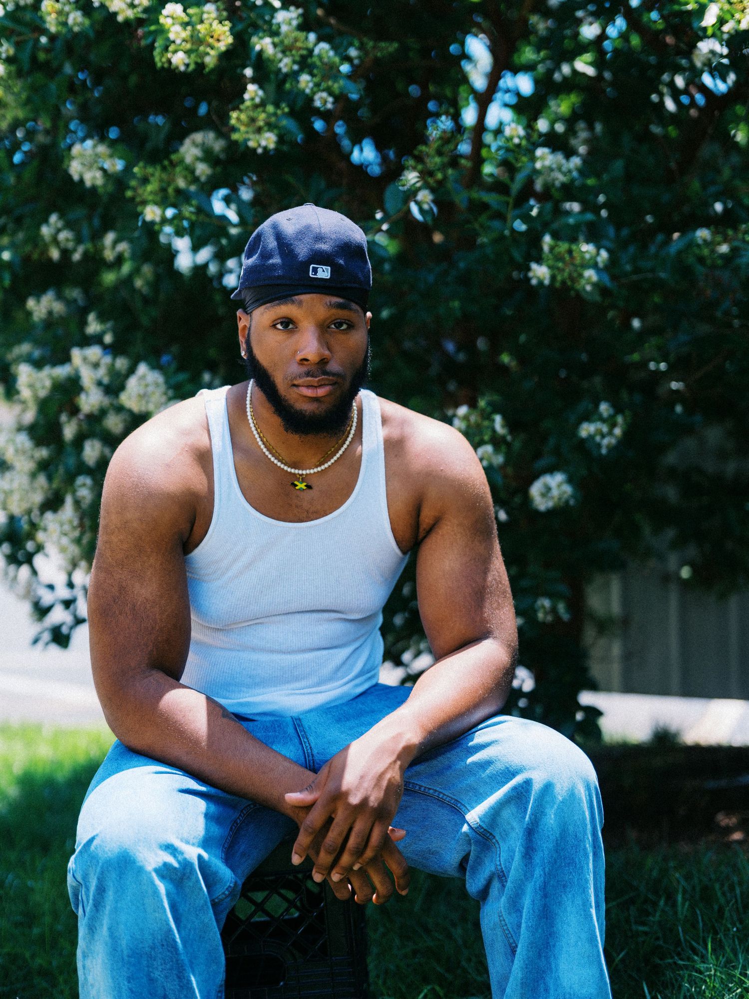 Young man in white tank top, blue jeans, and navy cap sits outdoors near flowering trees on green grass.