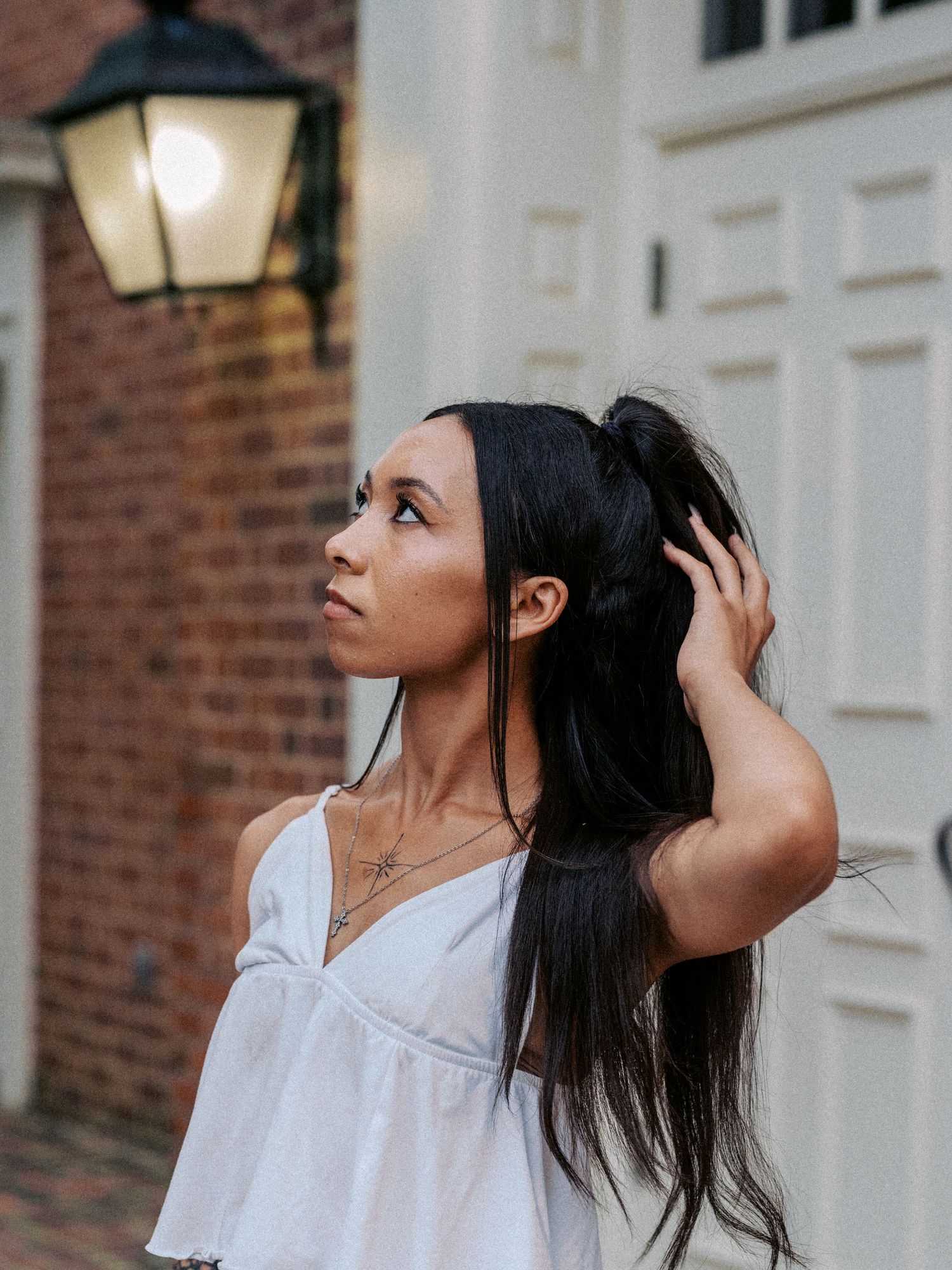 Young woman with long black hair in ponytail wearing white off-shoulder top, posing outdoors near brick wall and lantern.