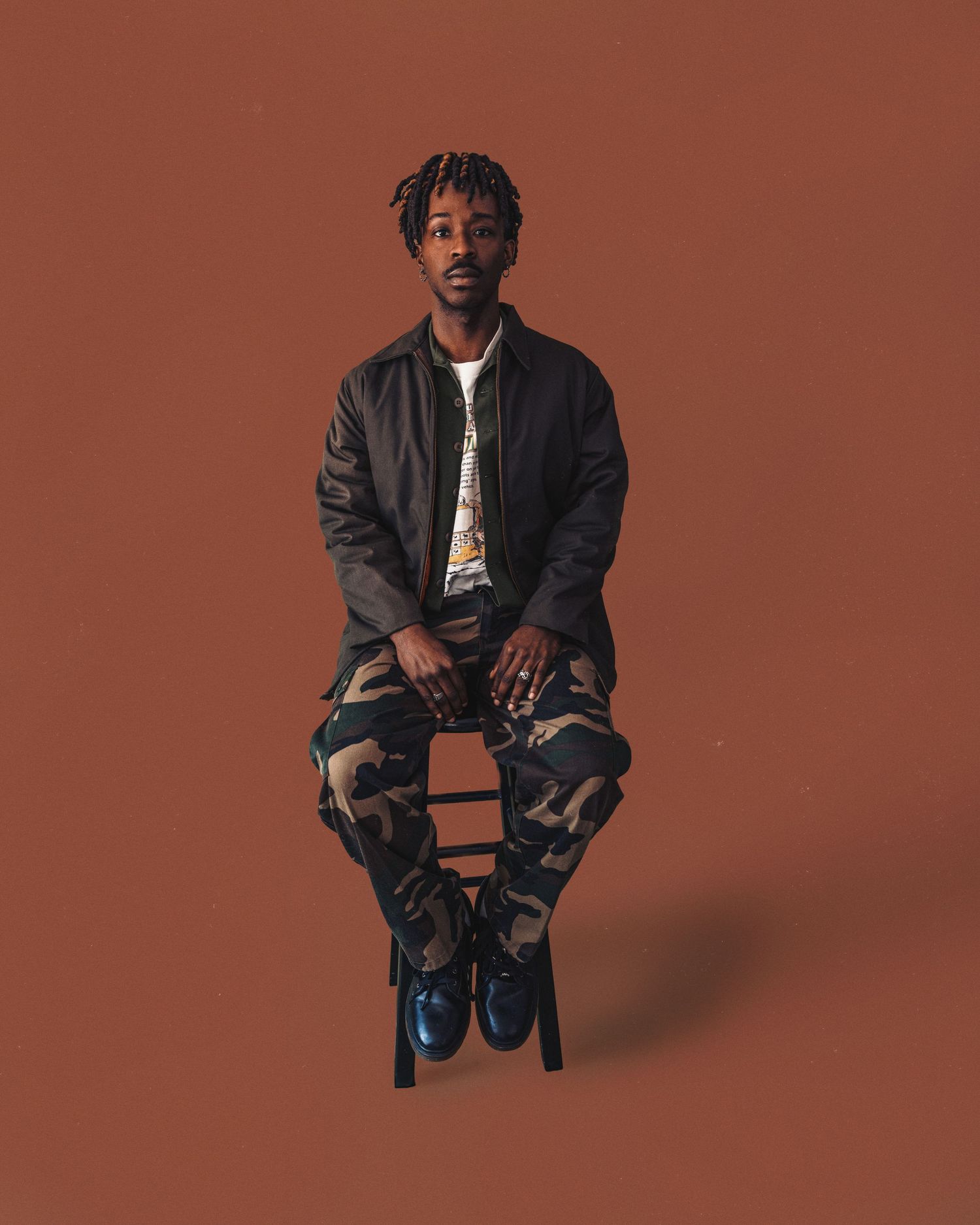 Young man with dreadlocks sits on stool wearing camo pants and dark jacket against brown background.