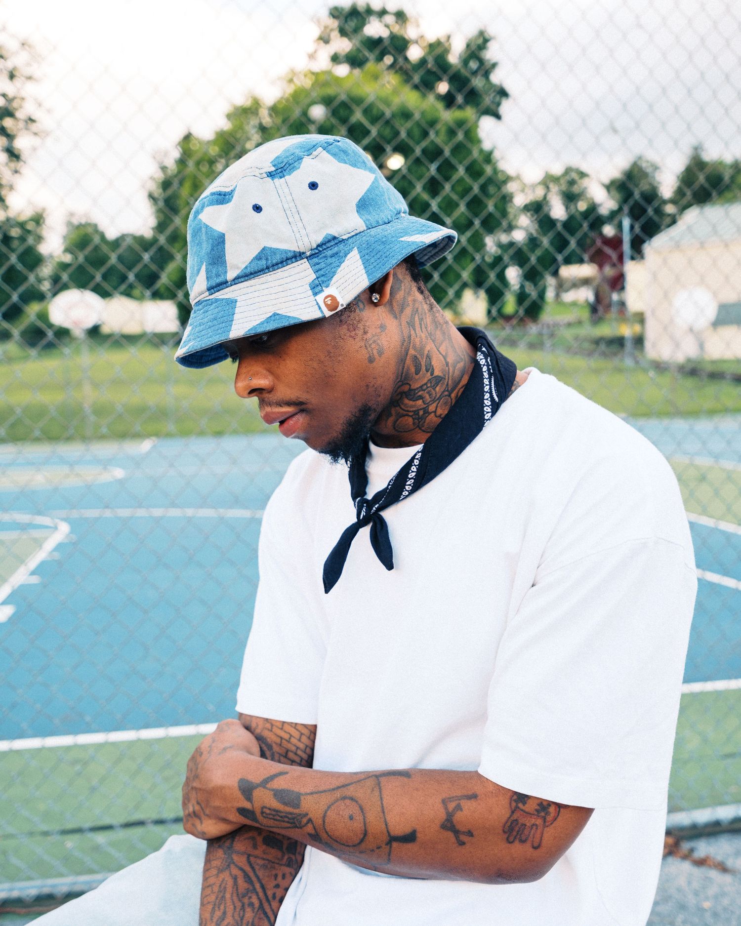 Tattooed man in white tee and blue star bucket hat stands by a tennis court fence, arms crossed.