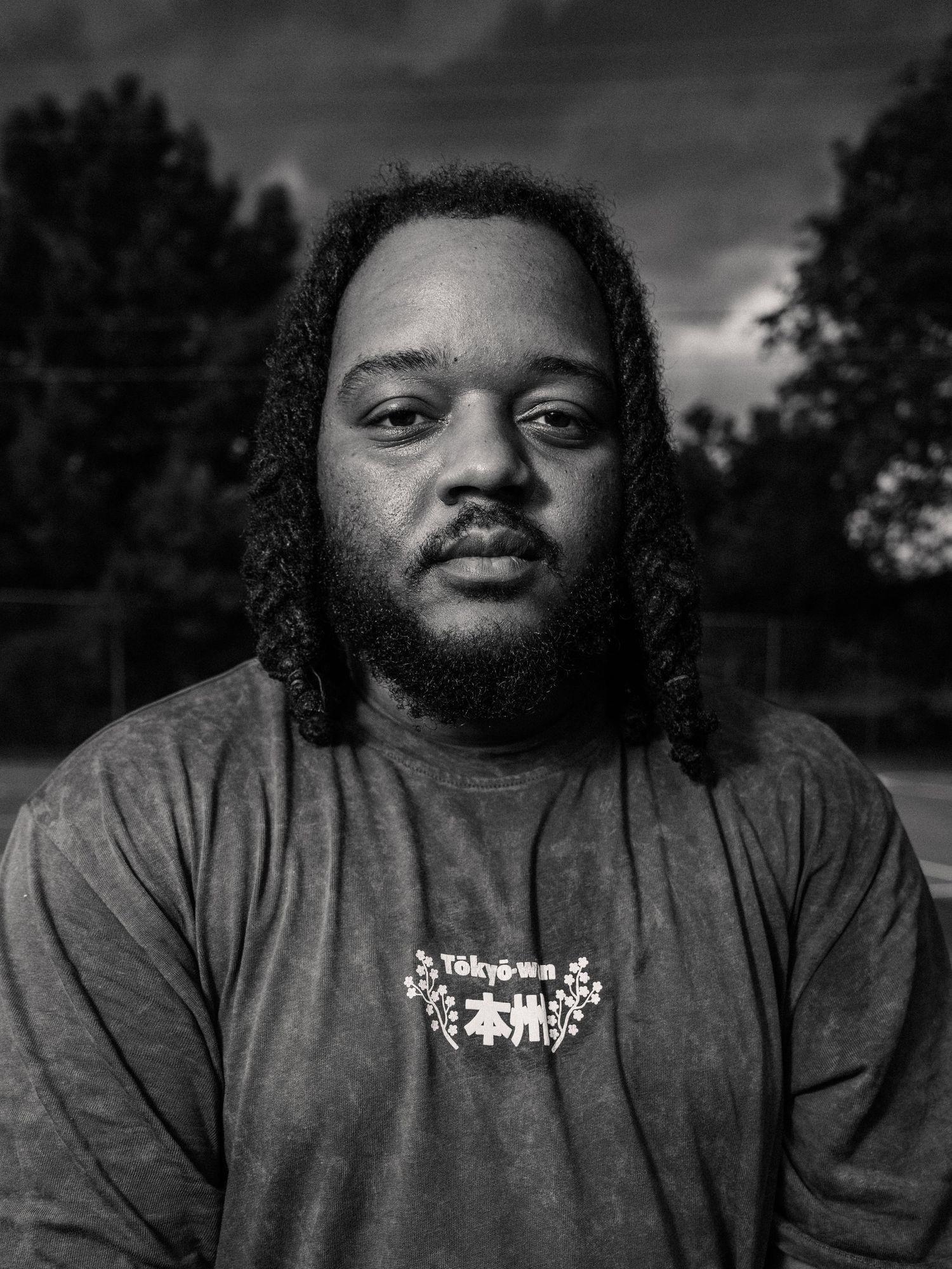 Black and white portrait of a bearded man with dreadlocks wearing a graphic t-shirt outdoors.