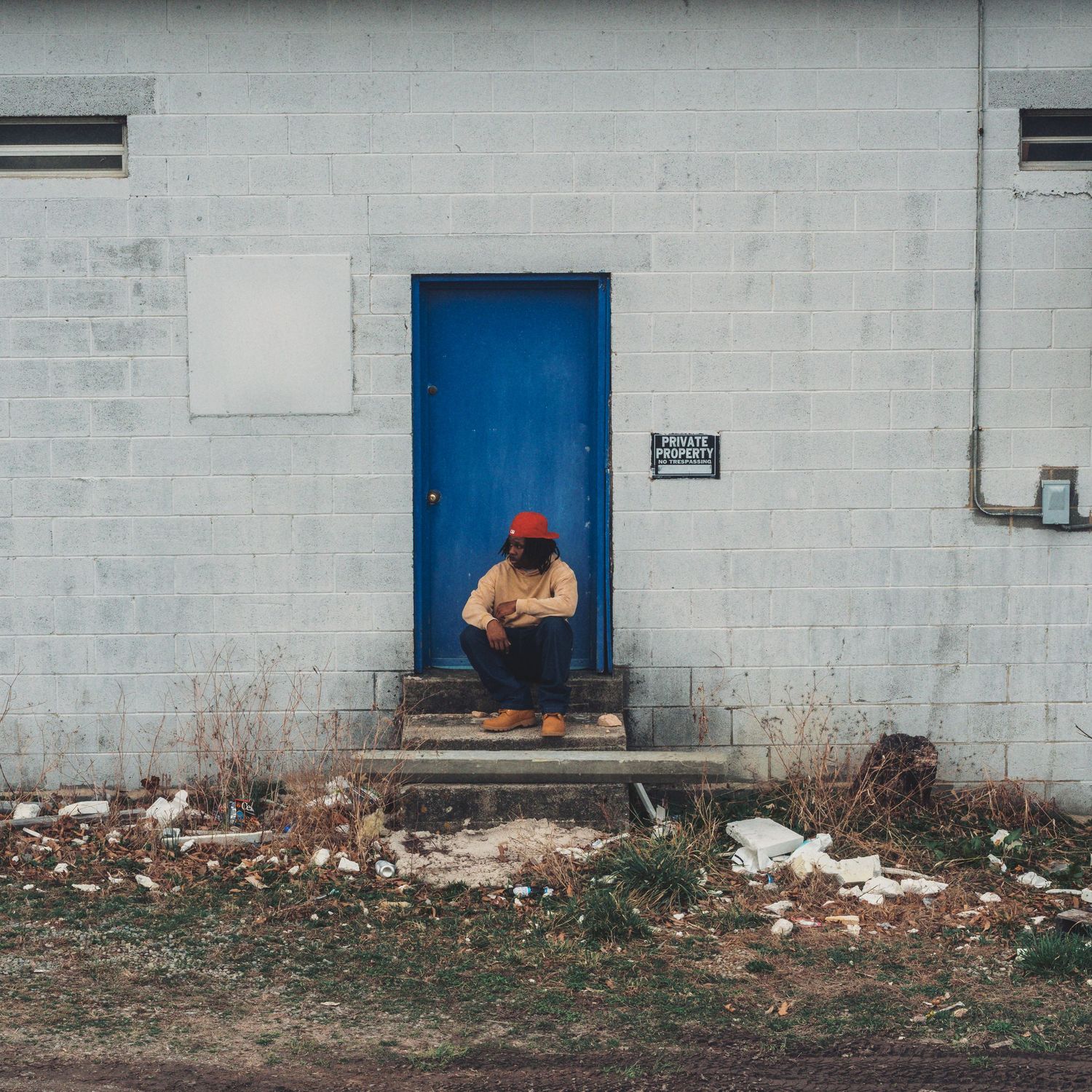 Person in red hat sitting on steps of a rundown building with a blue door and white brick wall.