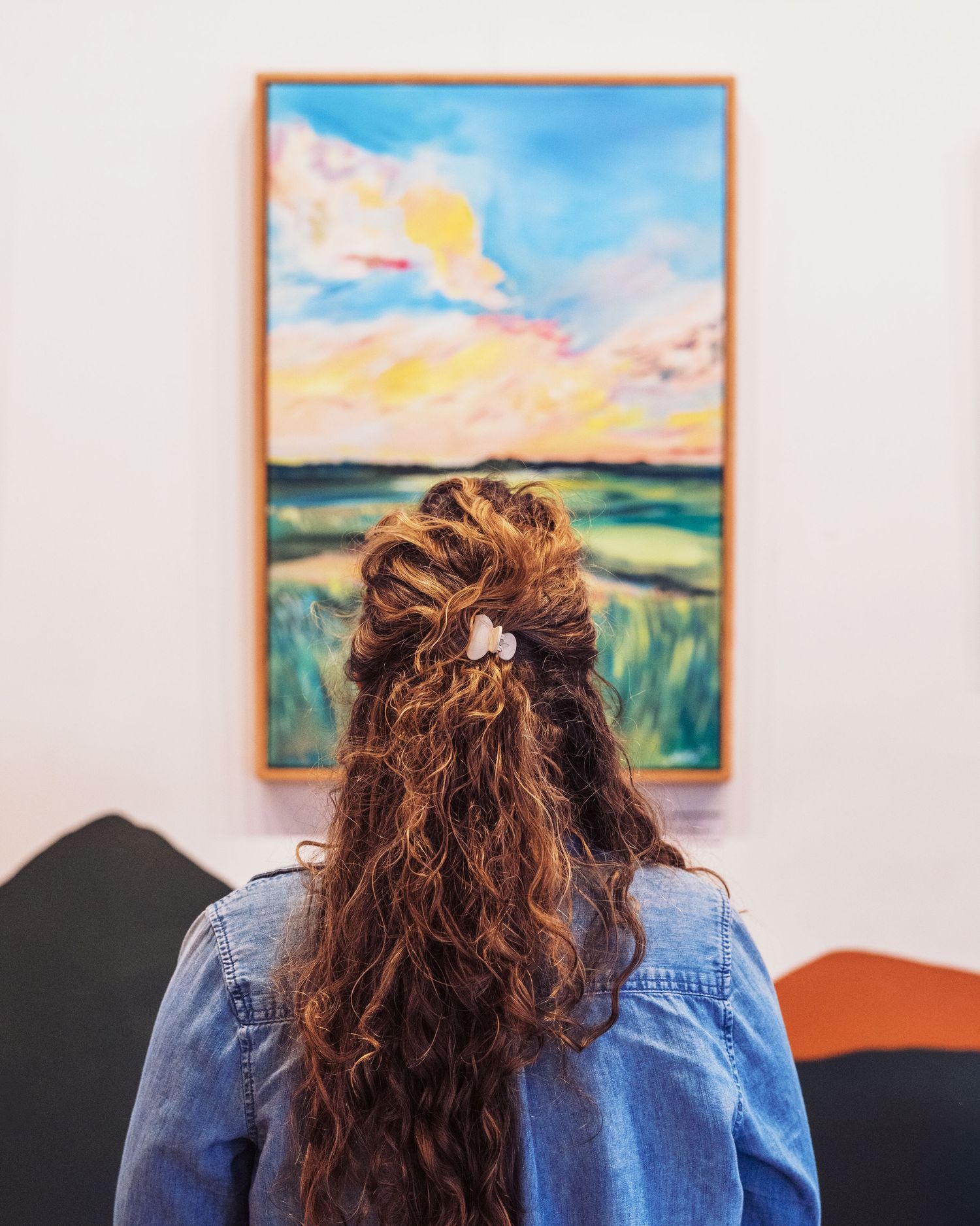 A woman with curly hair views a colorful landscape painting in an art gallery.