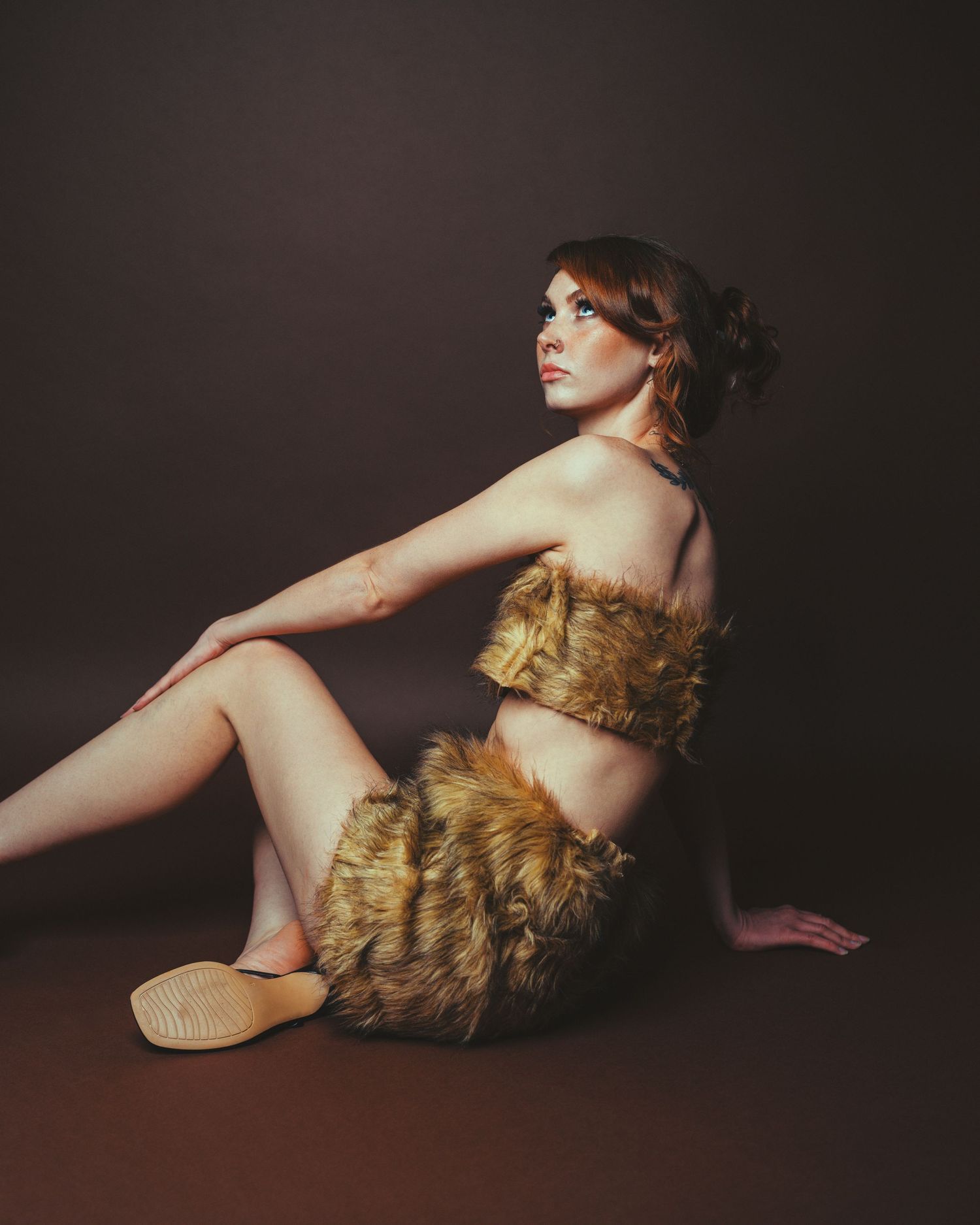 Woman in fur two-piece outfit seated on dark studio floor, posing elegantly with upswept red hair.