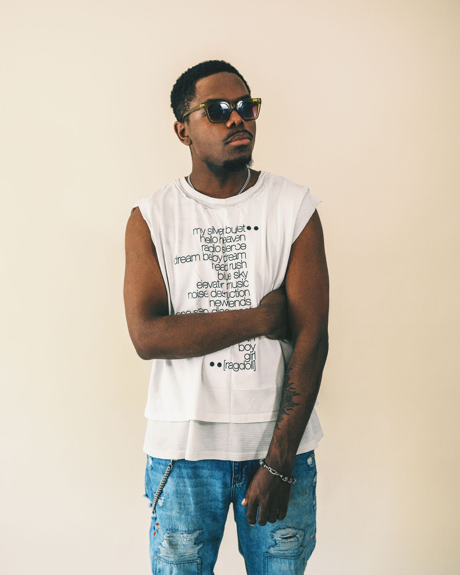 Young man in sunglasses wearing a white sleeveless graphic tee and ripped jeans, posing confidently against a beige wall.
