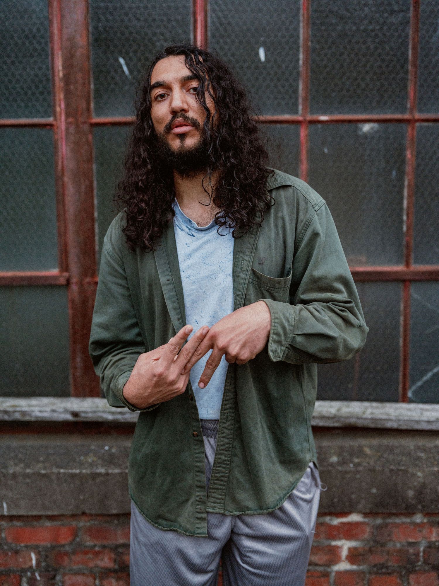 Person in olive jacket and gray shirt poses against brick wall in moody lighting.