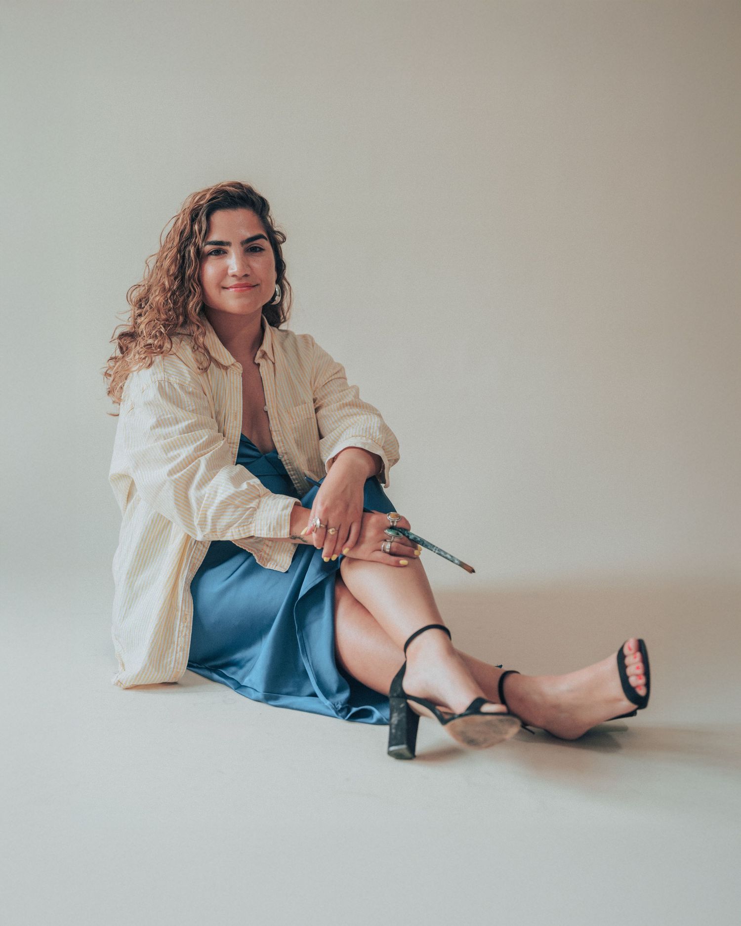 Fashion shoot of someone in cream blazer and blue dress lounging on floor against neutral backdrop.