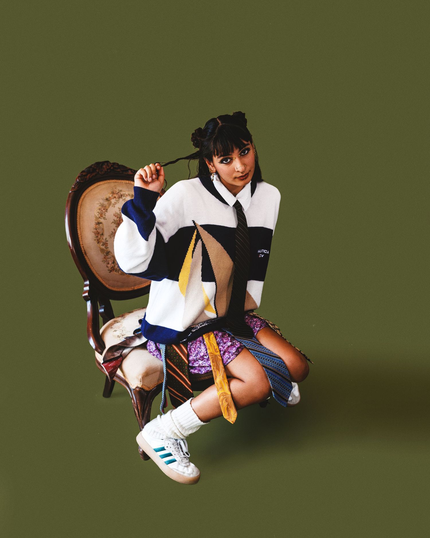 Young woman in white striped oversized jacket, purple skirt, and Adidas sneakers posing on vintage chair against olive background.