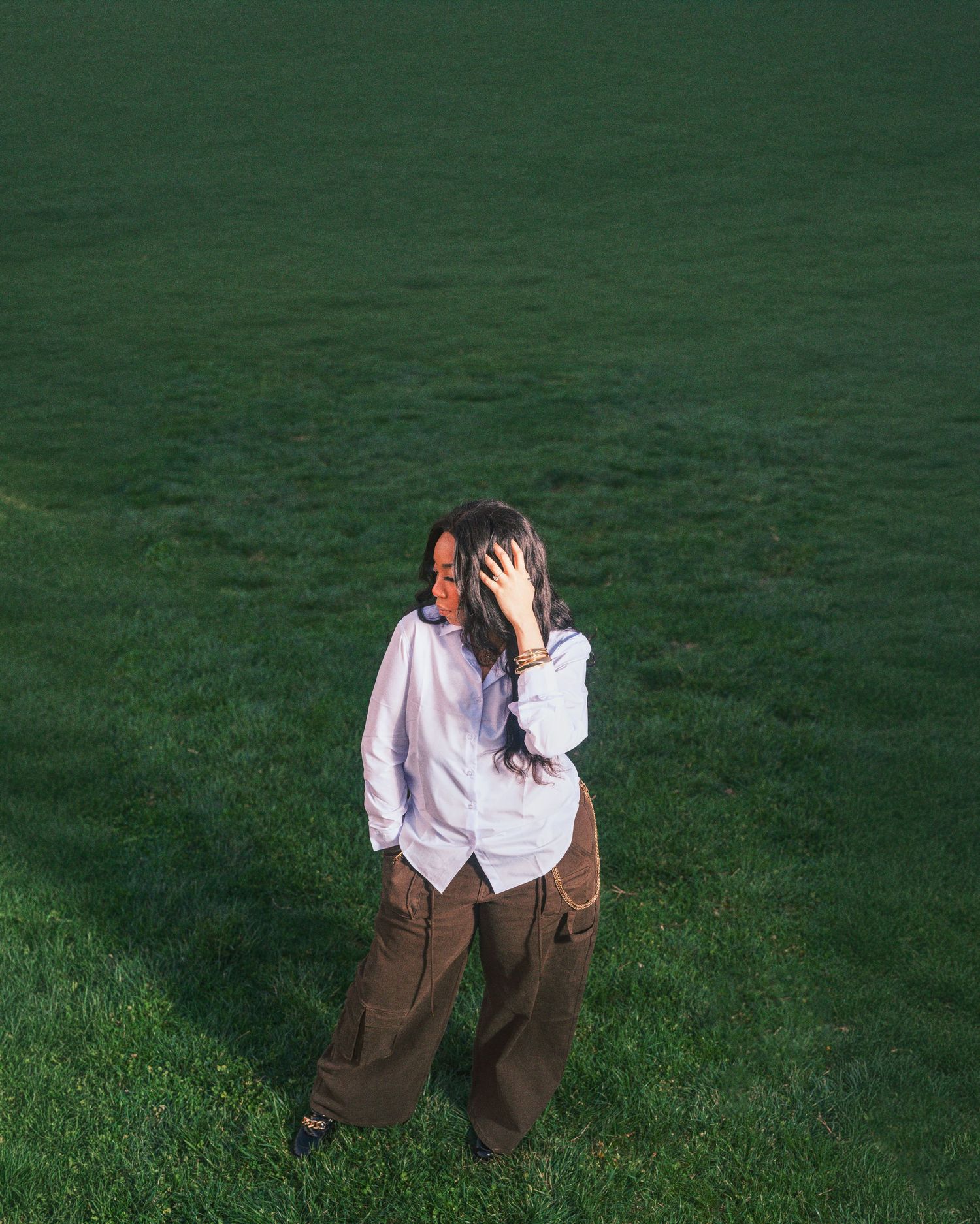 Woman in white shirt and brown cargo pants standing on a lush green grass field in golden hour light.