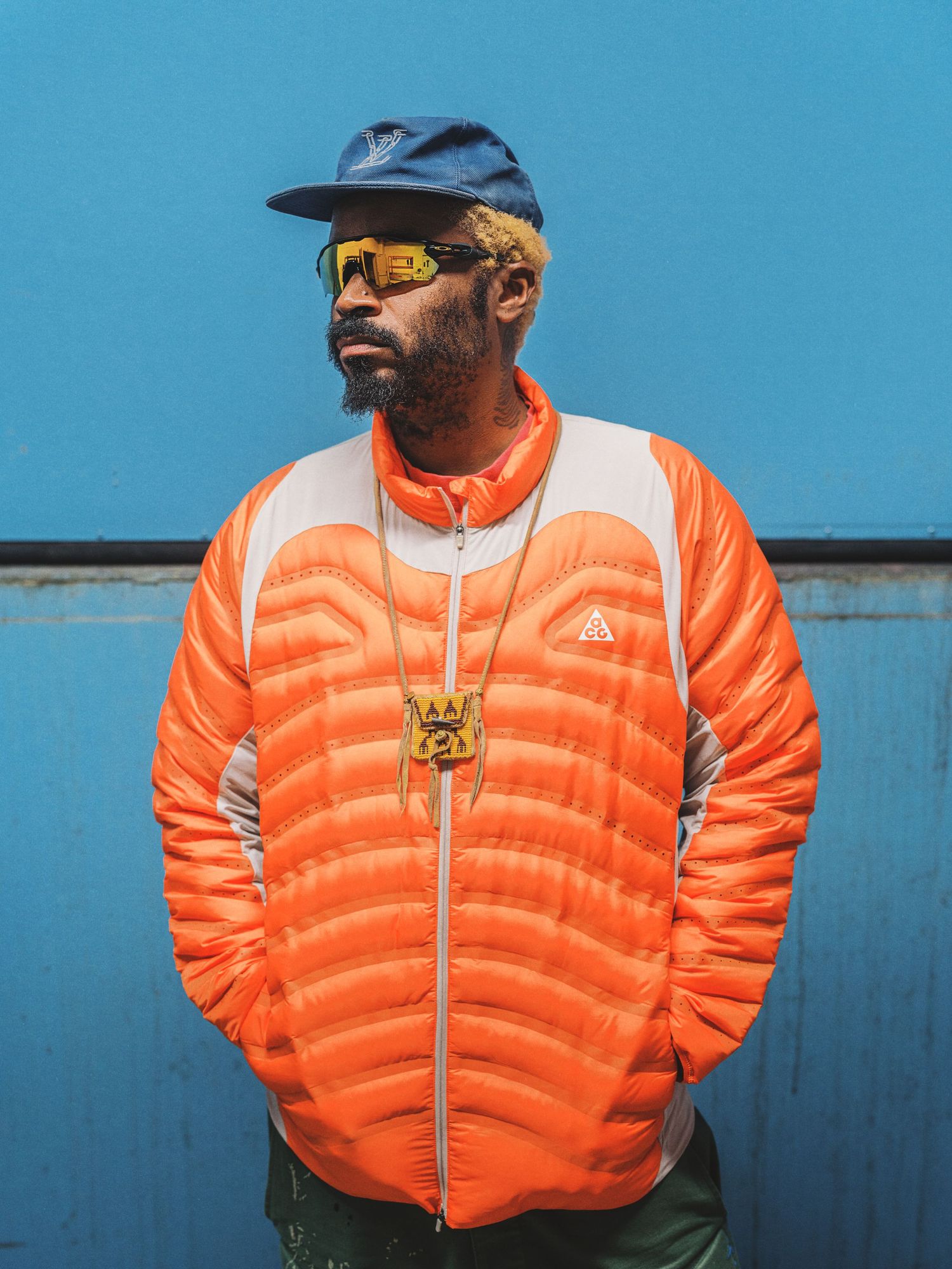 Man wearing orange Nike ACG puffer jacket, blue cap, and yellow sunglasses standing against a teal wall.