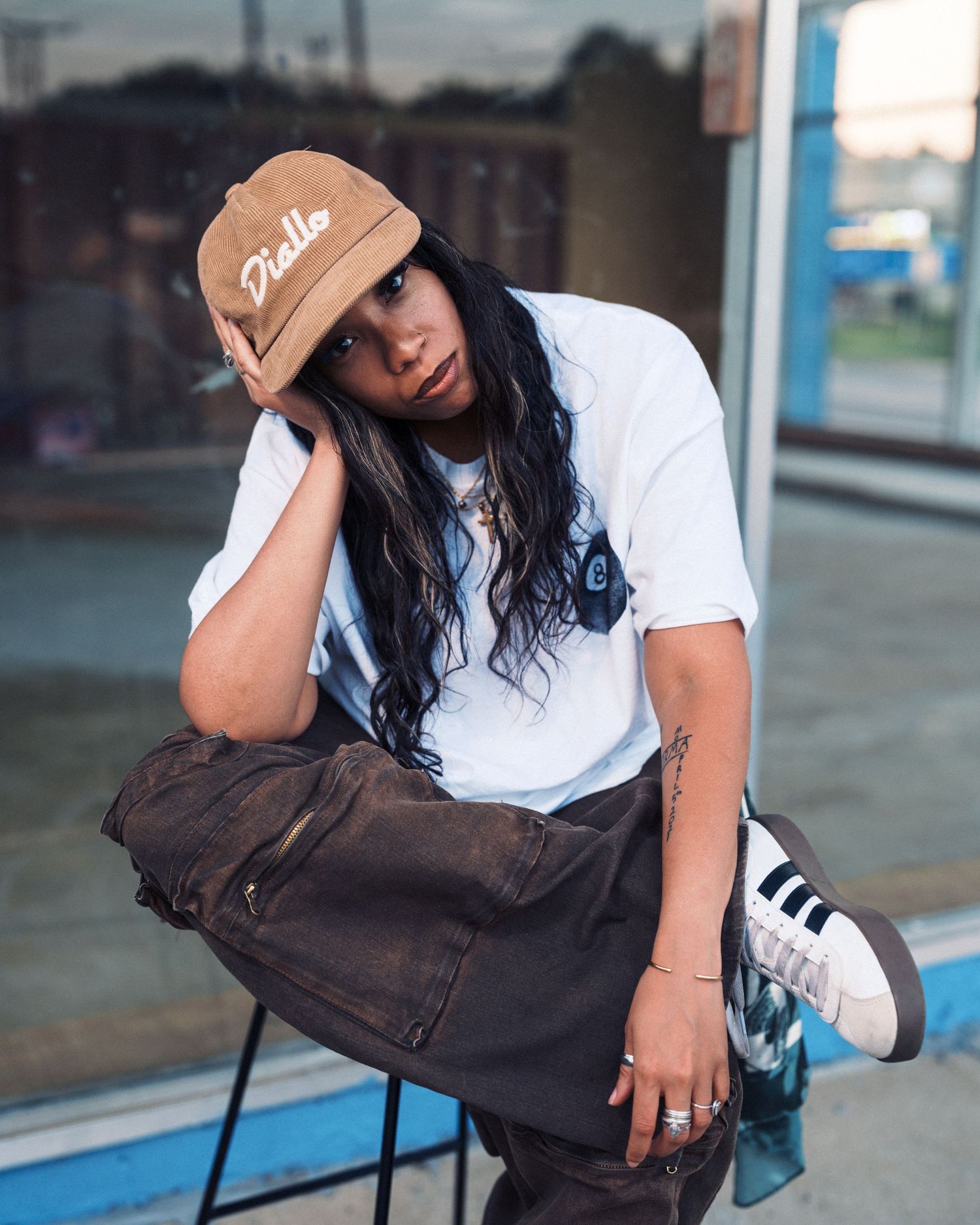 Young woman in white tee, brown cap, dark pants, and Adidas sneakers sitting casually outside a storefront.