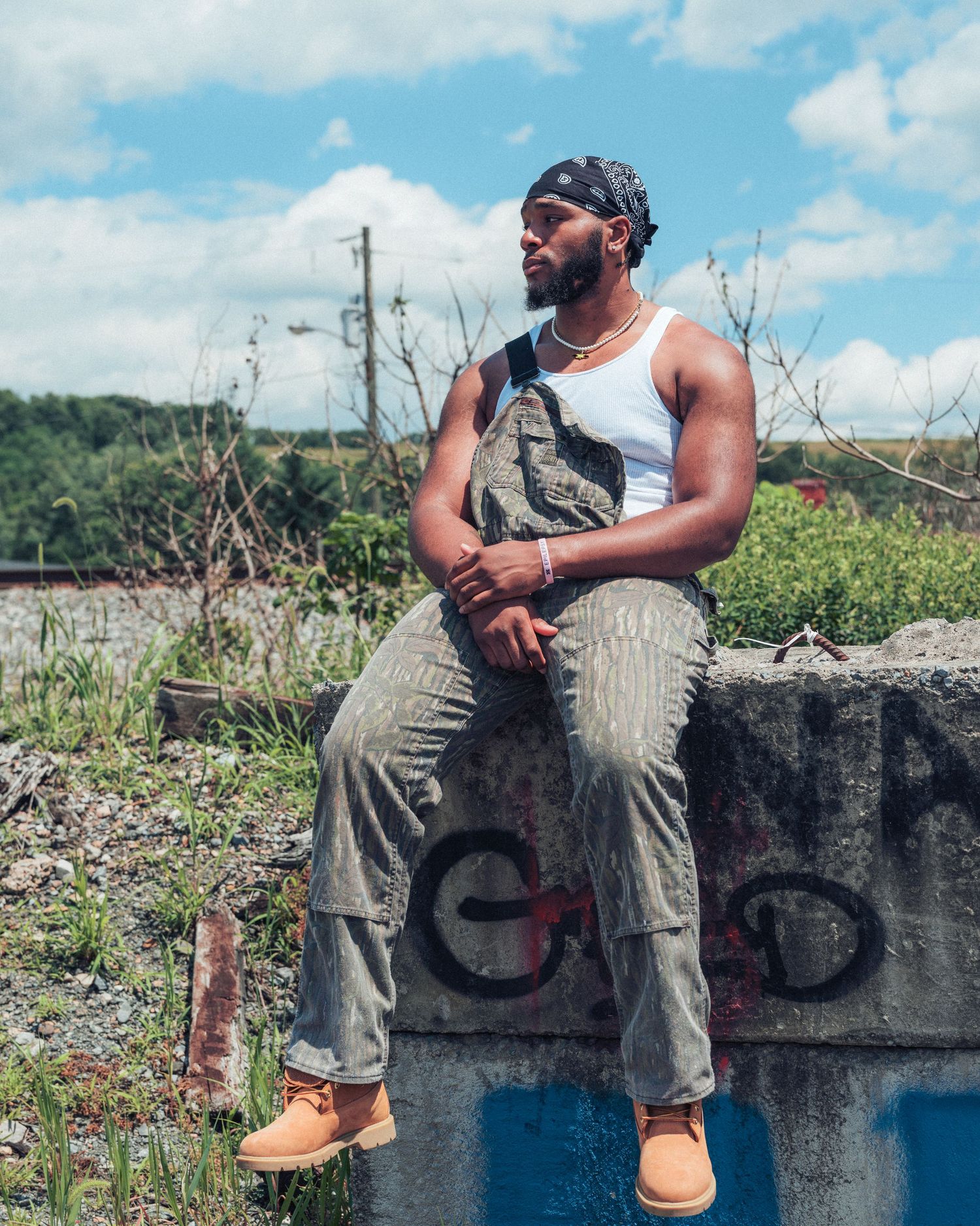 Man in overalls and bandana sits on graffiti concrete wall outdoors on cloudy day.