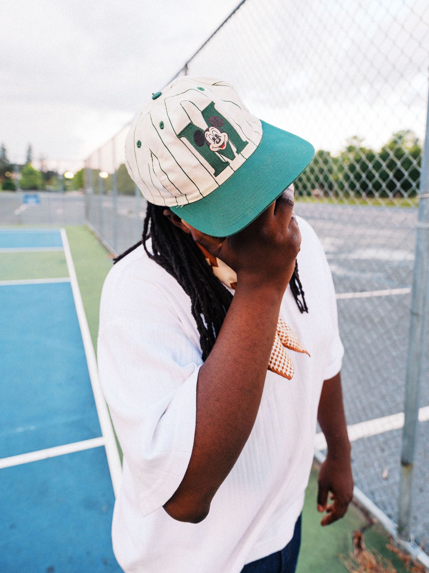Young man with dreadlocks wearing a white pinstripe snapback cap and white tee near a chain-link fence on a tennis court.