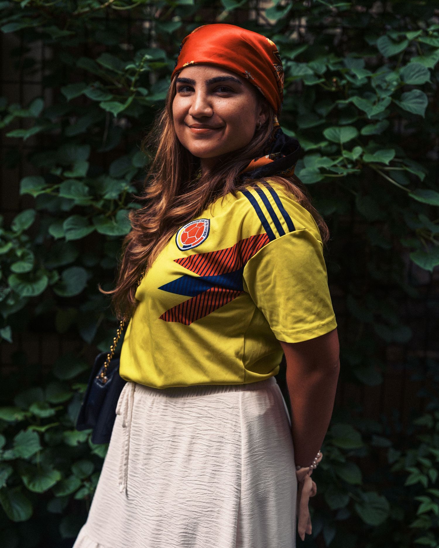 Woman smiling in yellow Colombia soccer jersey, orange headband, white skirt, posing outdoors against green leafy background.