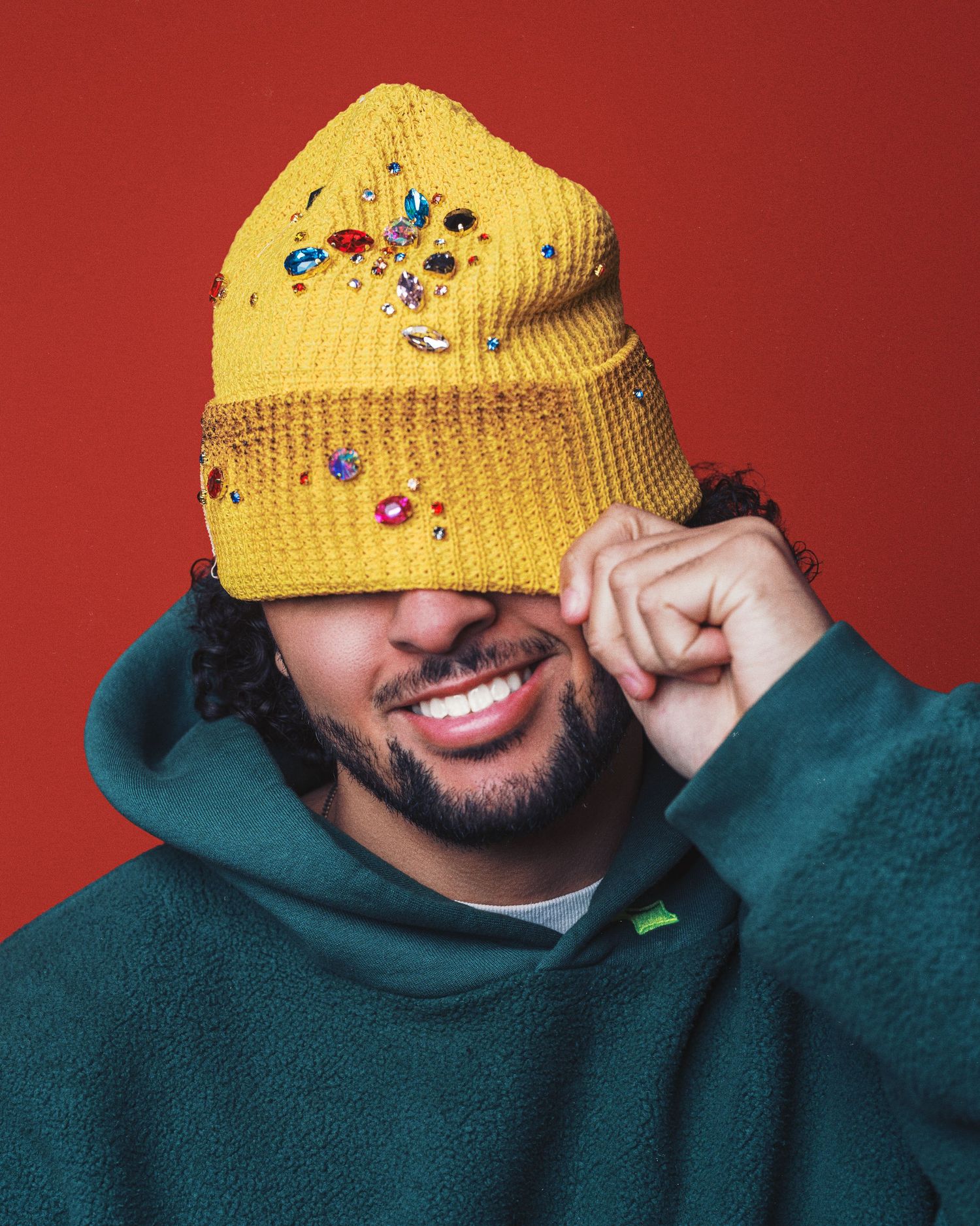 A person in a green hoodie and yellow beaded beanie smiles while touching the brim of their hat against a red background.