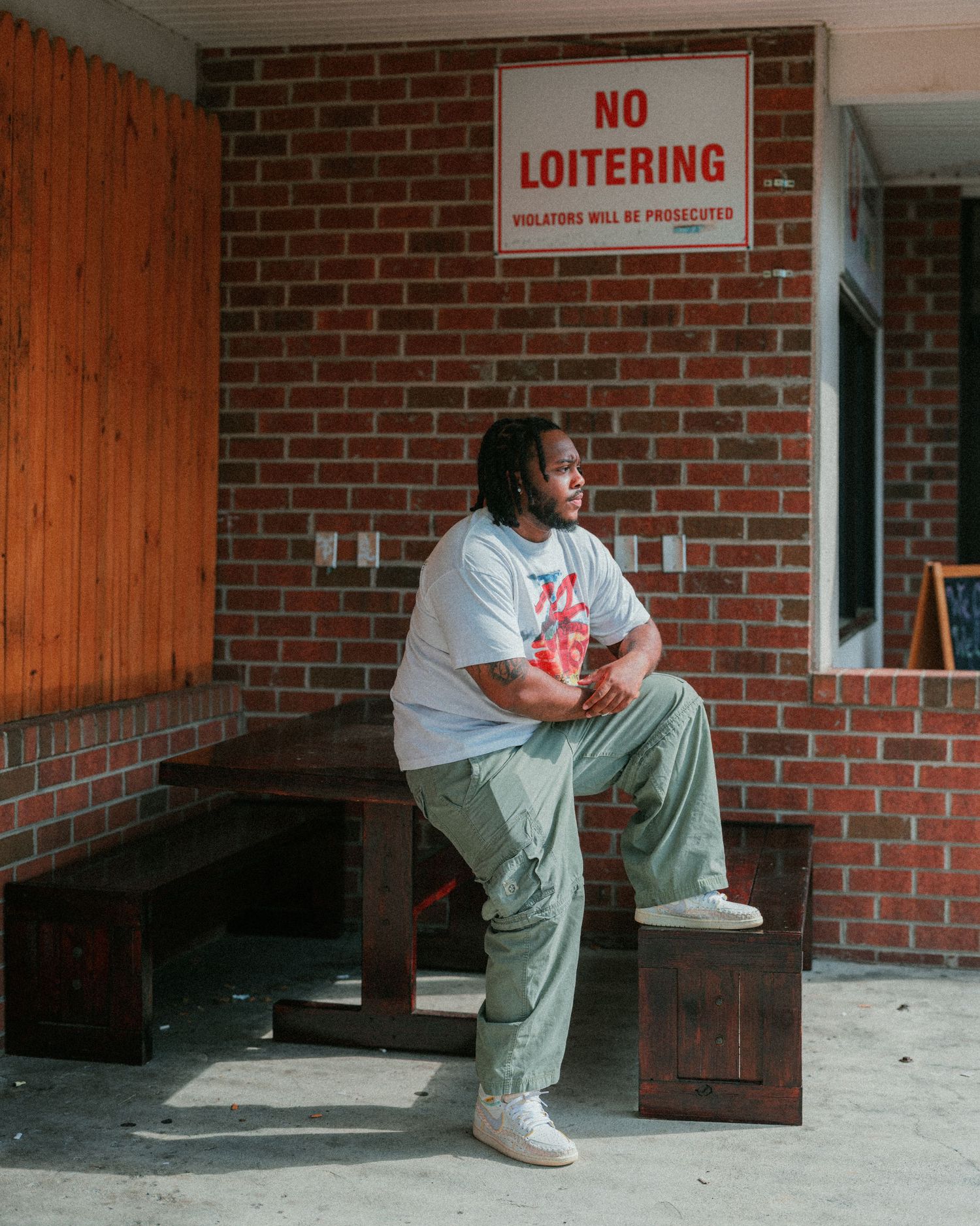 Person sitting on a wooden bench under a No Loitering sign on a brick wall exterior.