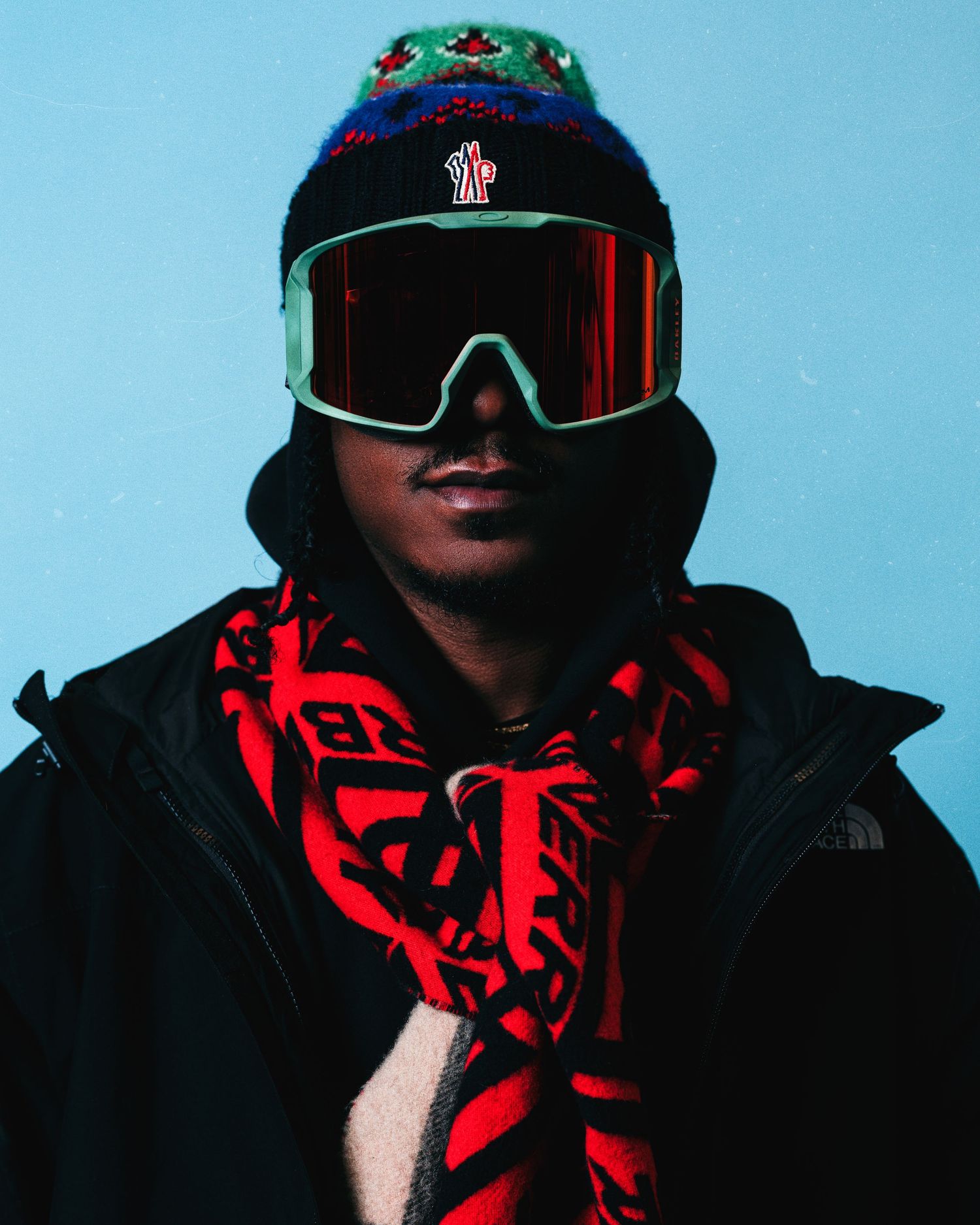 Person wearing ski goggles, colorful beanie, red patterned scarf, and black North Face jacket against blue background.
