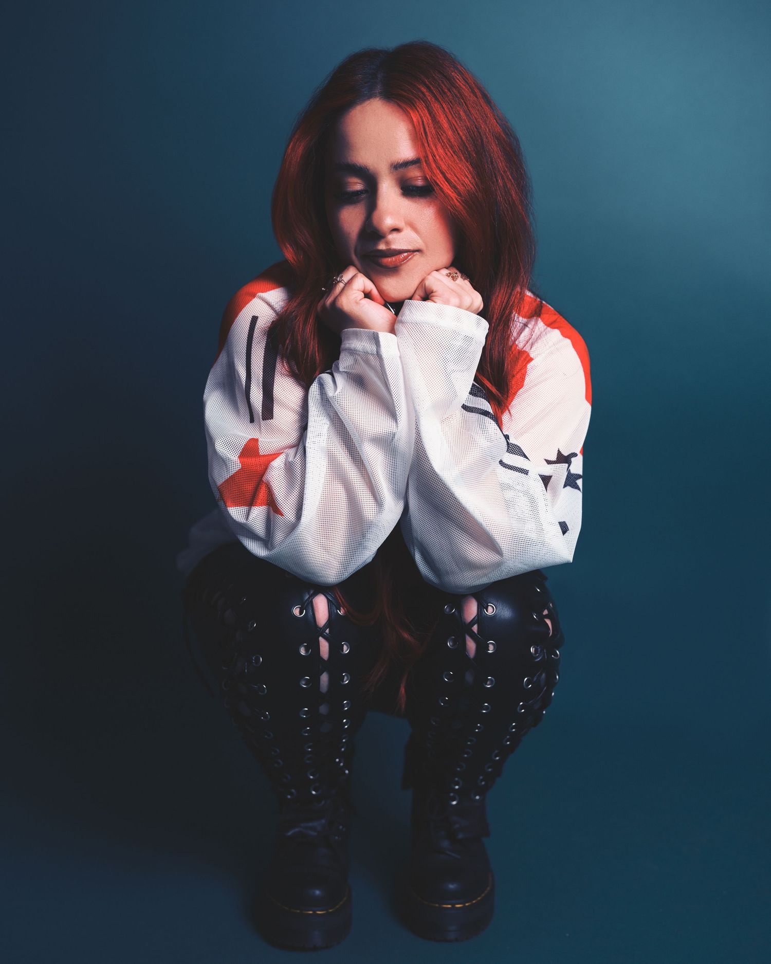Red-haired woman in white jacket and black lace-up boots poses thoughtfully against a teal background.