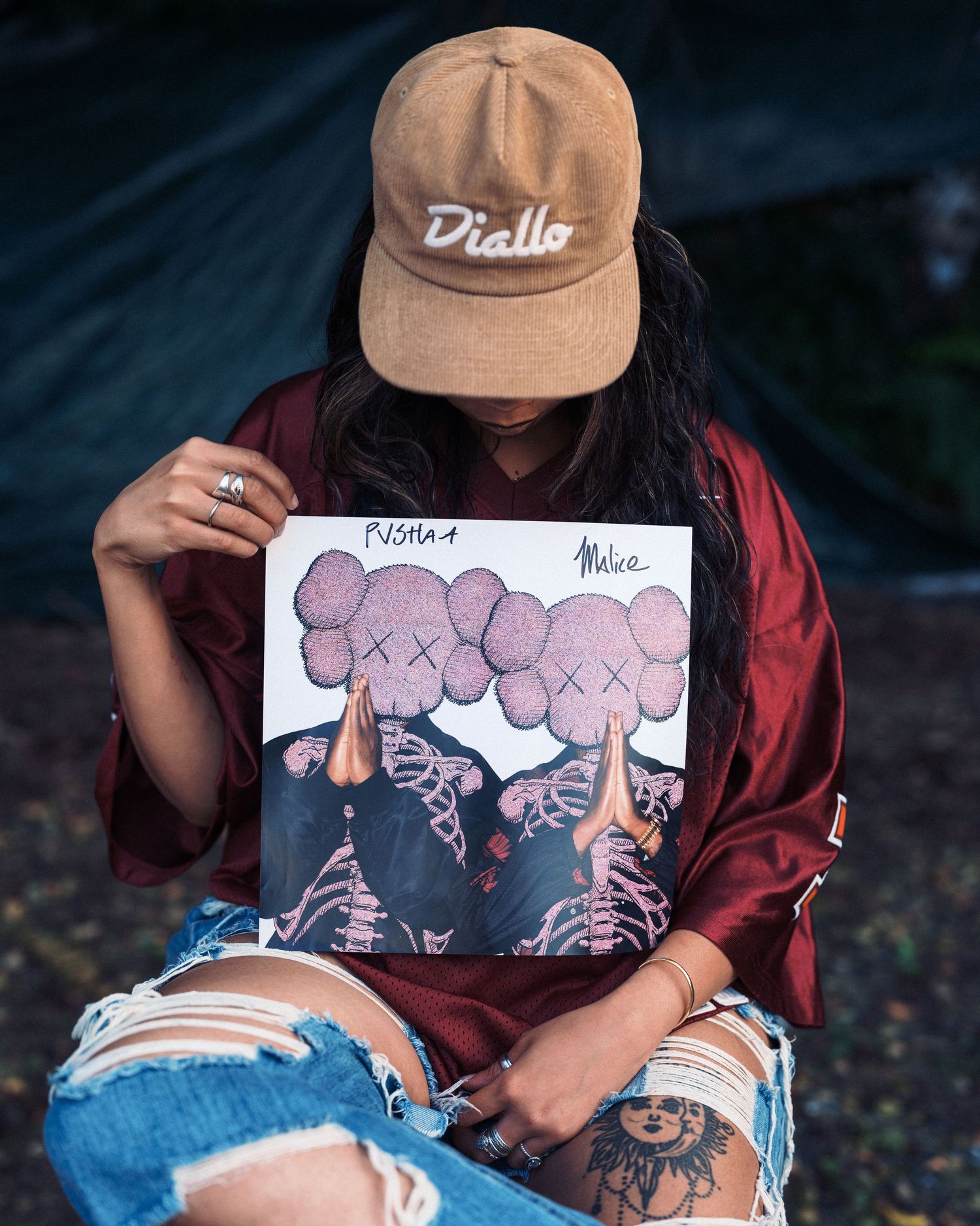 Person holding KAWS vinyl record album wearing a Diallo cap and ripped jeans outdoors.