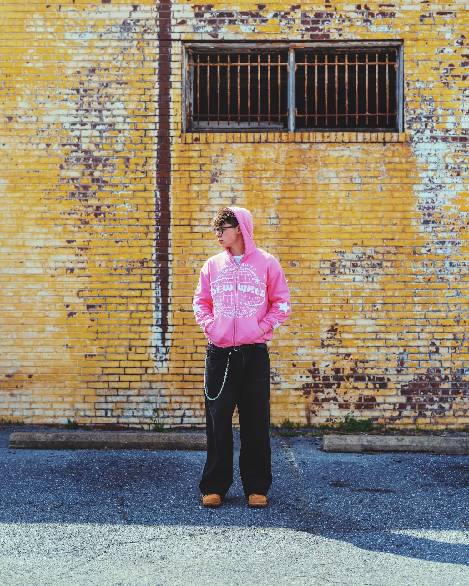 Person in pink hoodie and black wide-leg pants standing against a yellow brick wall with a barred window.