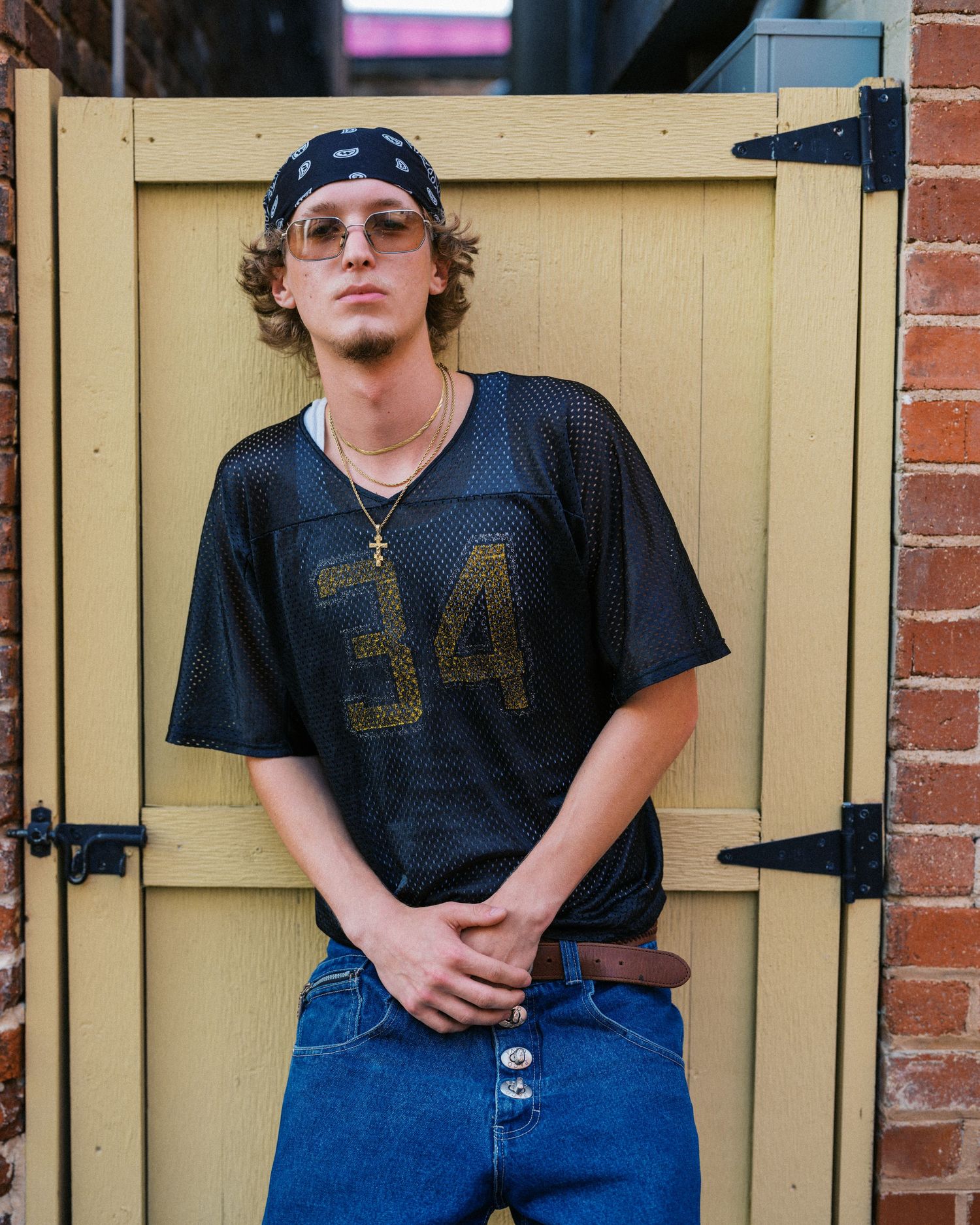 Young man in black mesh jersey number 34, jeans, bandana, and tinted glasses leaning against a yellow wooden door.