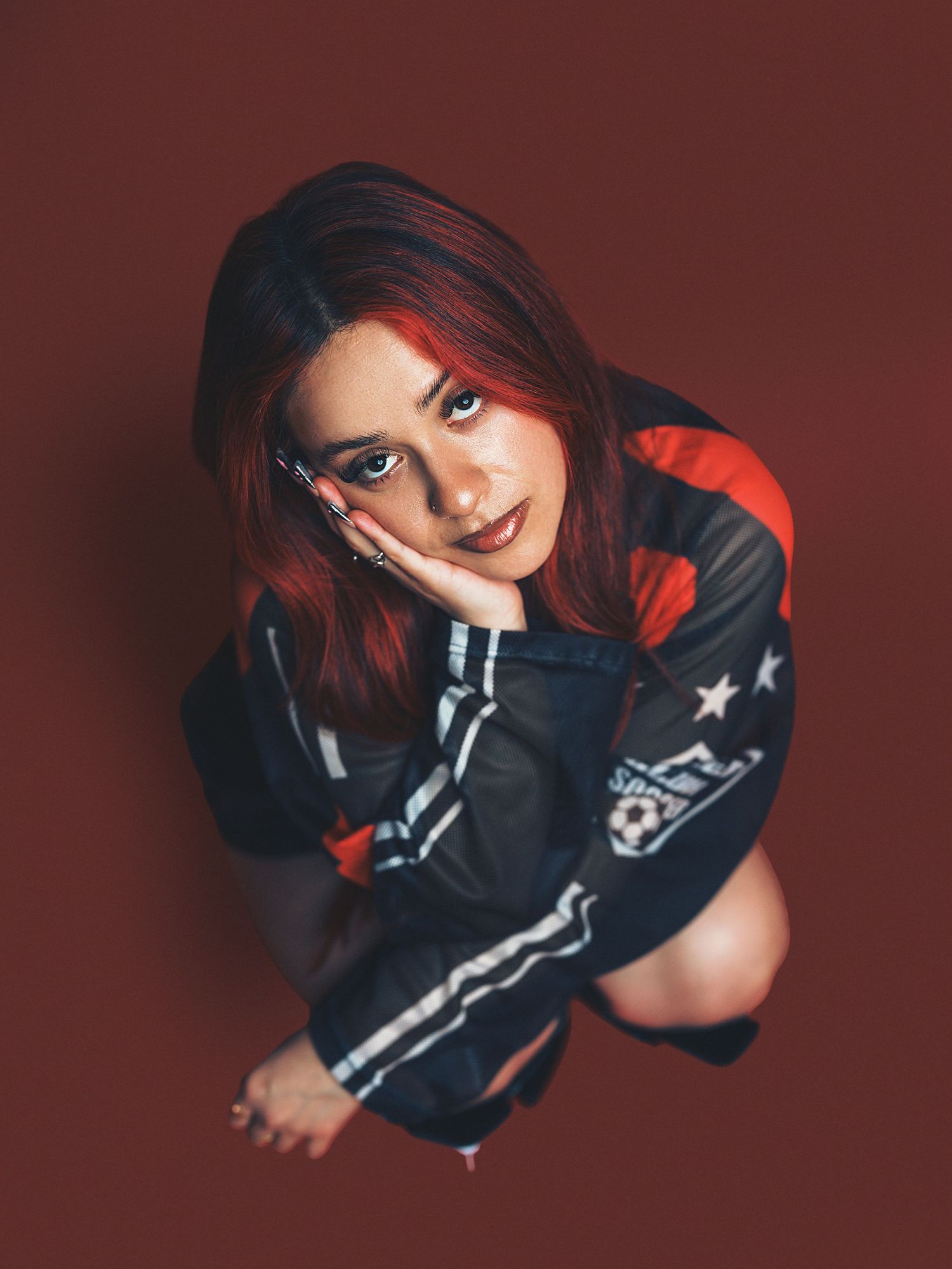 Young woman with red hair in a sports jacket poses against a dark red background, looking up at the camera.