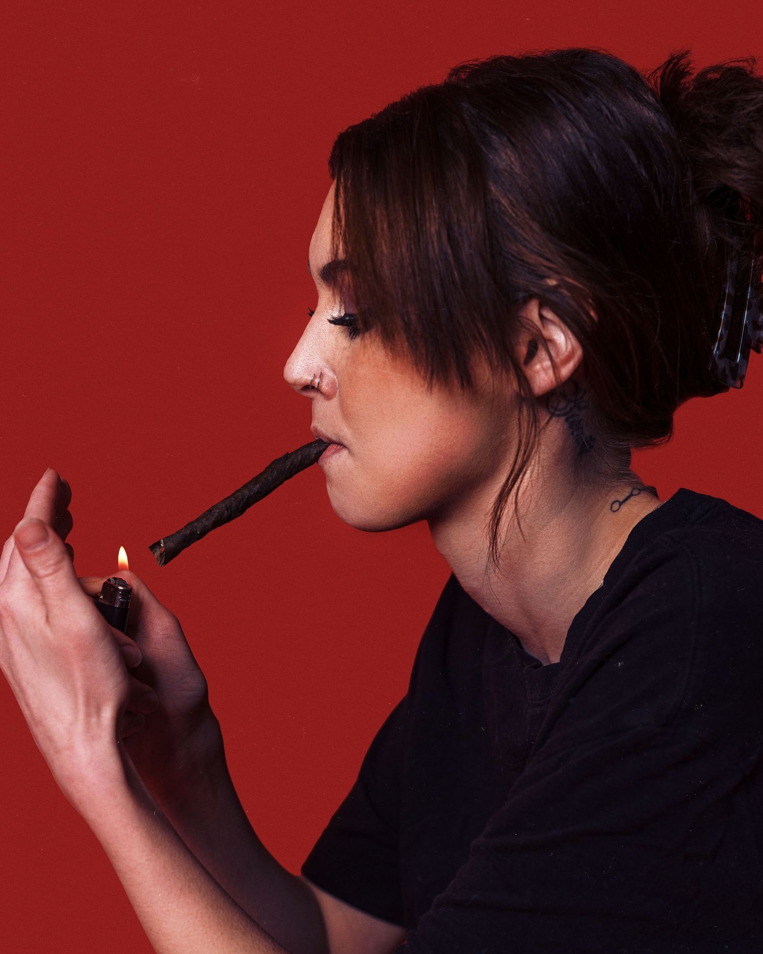Young woman with updo hairstyle lighting a cigarette against a bold red background.