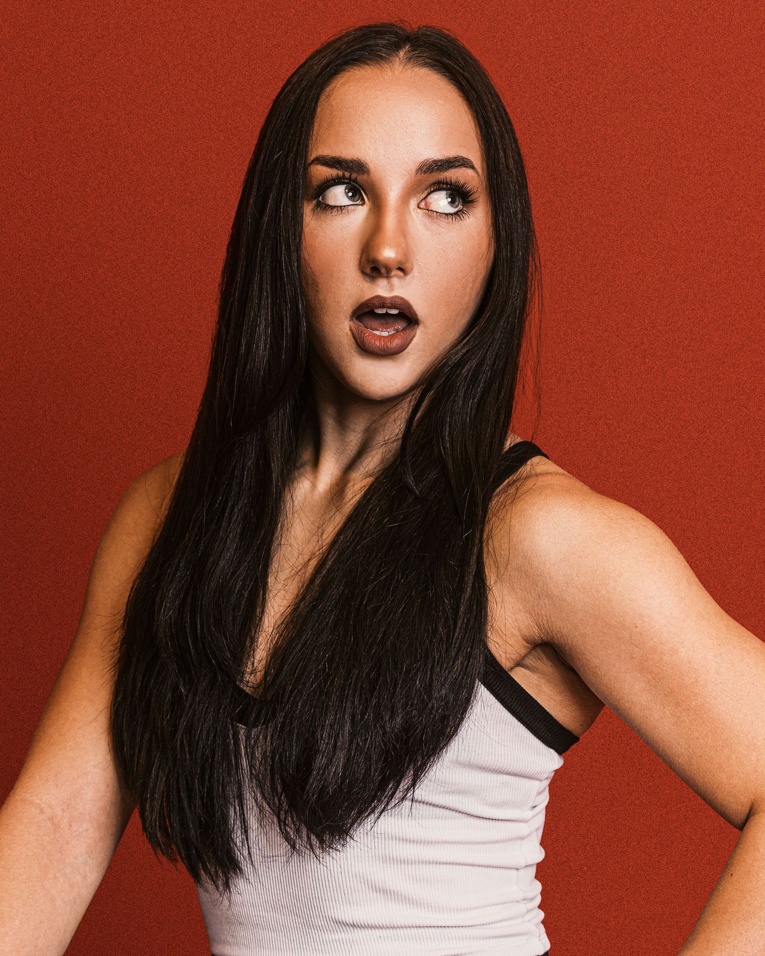 A woman with dark hair and bold red lipstick poses expressively against a red background in a white tank top.
