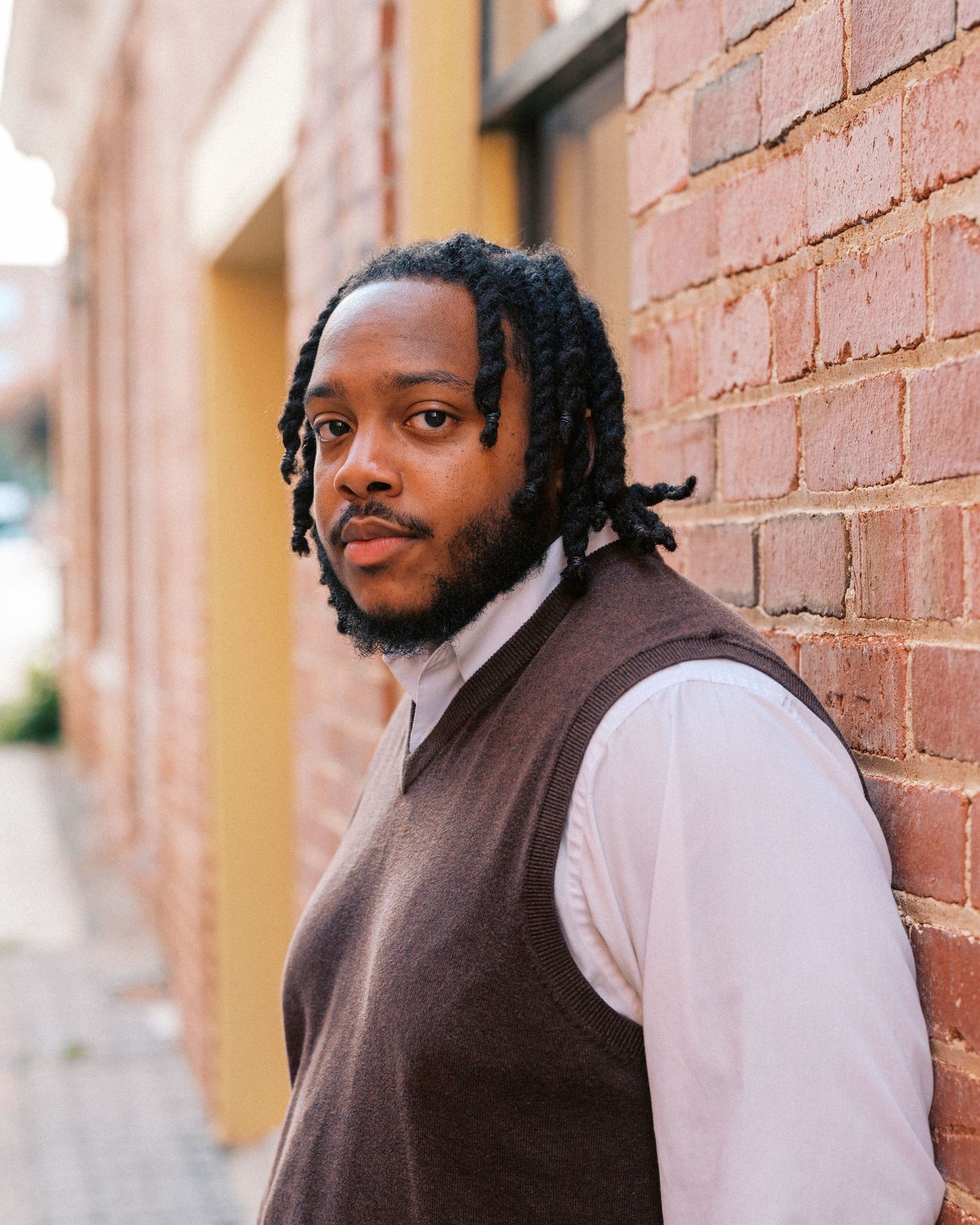 Young Black man with dreadlocks wearing a brown vest over white shirt, leaning against a red brick wall outdoors.