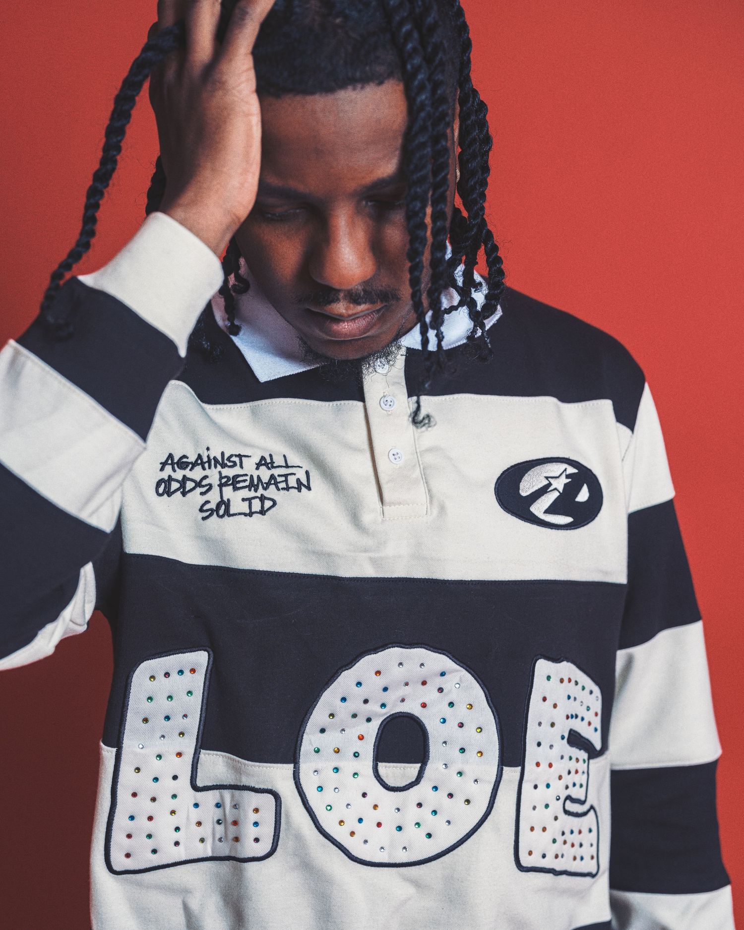 Young man in striped black and white LOL rugby shirt with braids poses against red background.