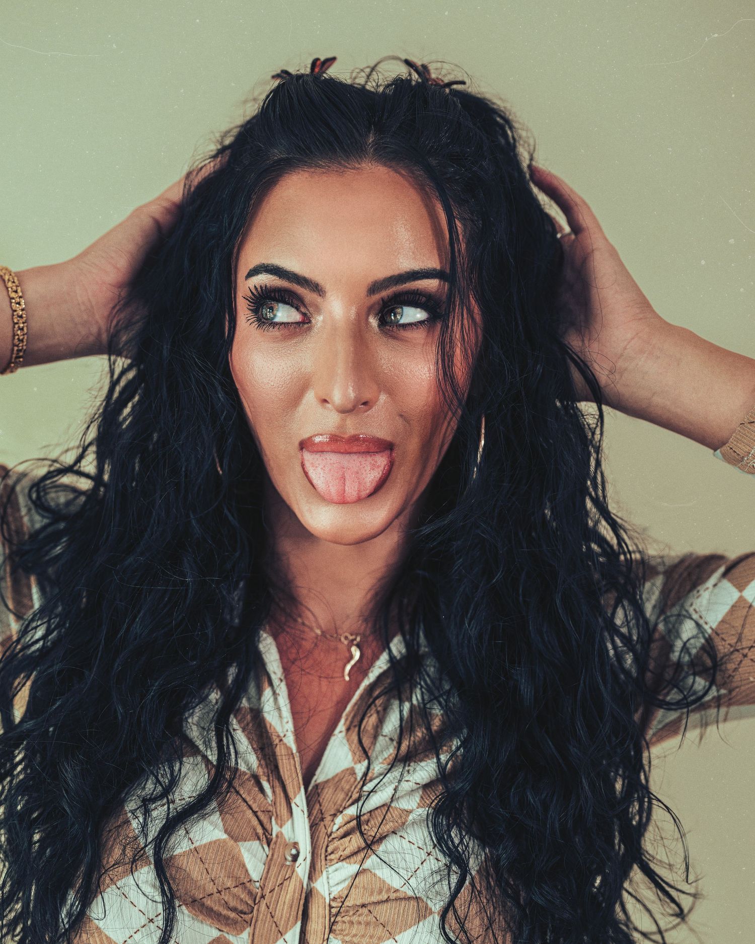 Woman with long dark wavy hair, bold makeup, sticking tongue out while holding hair with both hands.