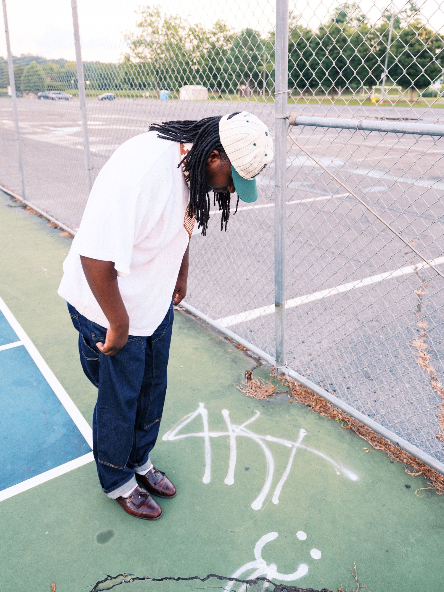 Person with dreadlocks in white tee and jeans stands by chain-link fence on outdoor basketball court with graffiti.