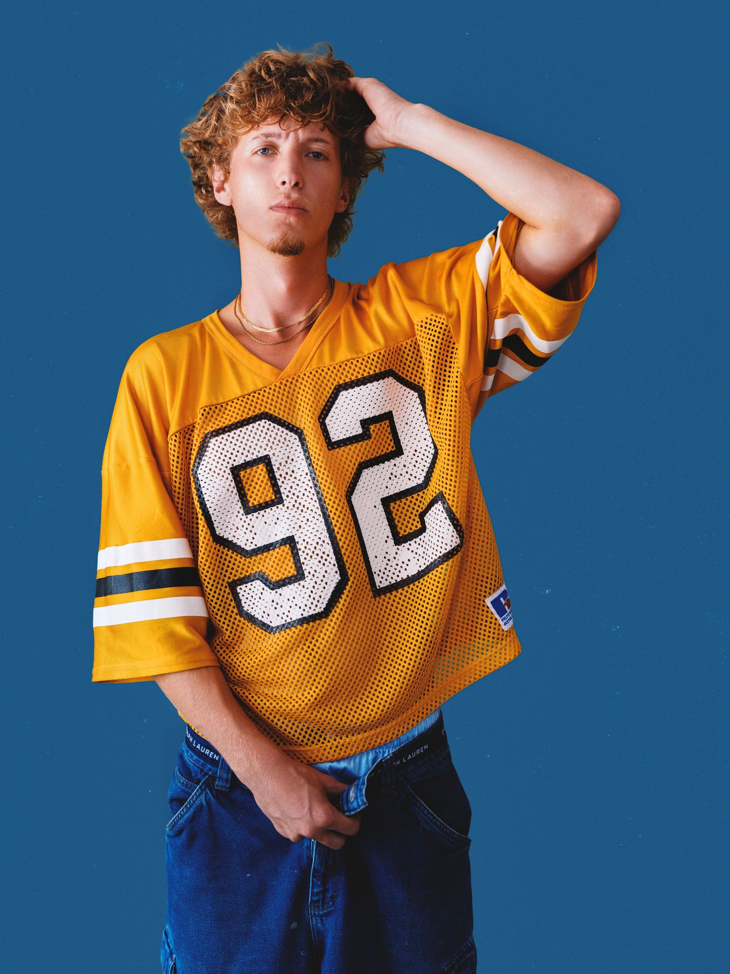 Young man in yellow number 92 football jersey and blue jeans posing against a blue background.