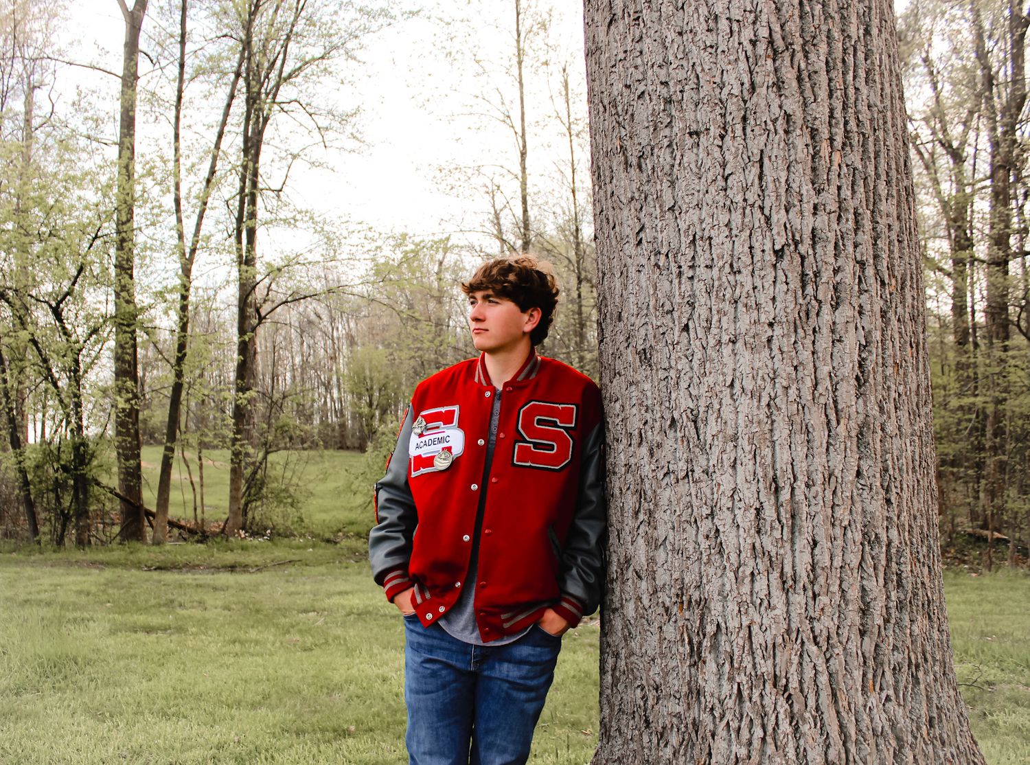 male high school senior stands leaning against a tree, looking off into the distance with a wooded area behind him in the background.