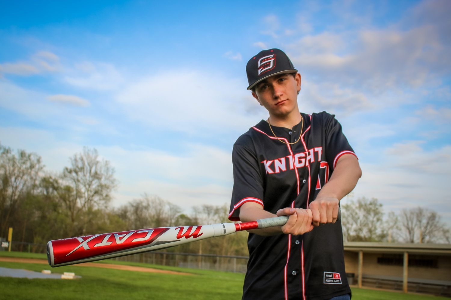 a male high school senior stands on baseball field and holds baseball bat in downswing position as he looks seriously at the camera.